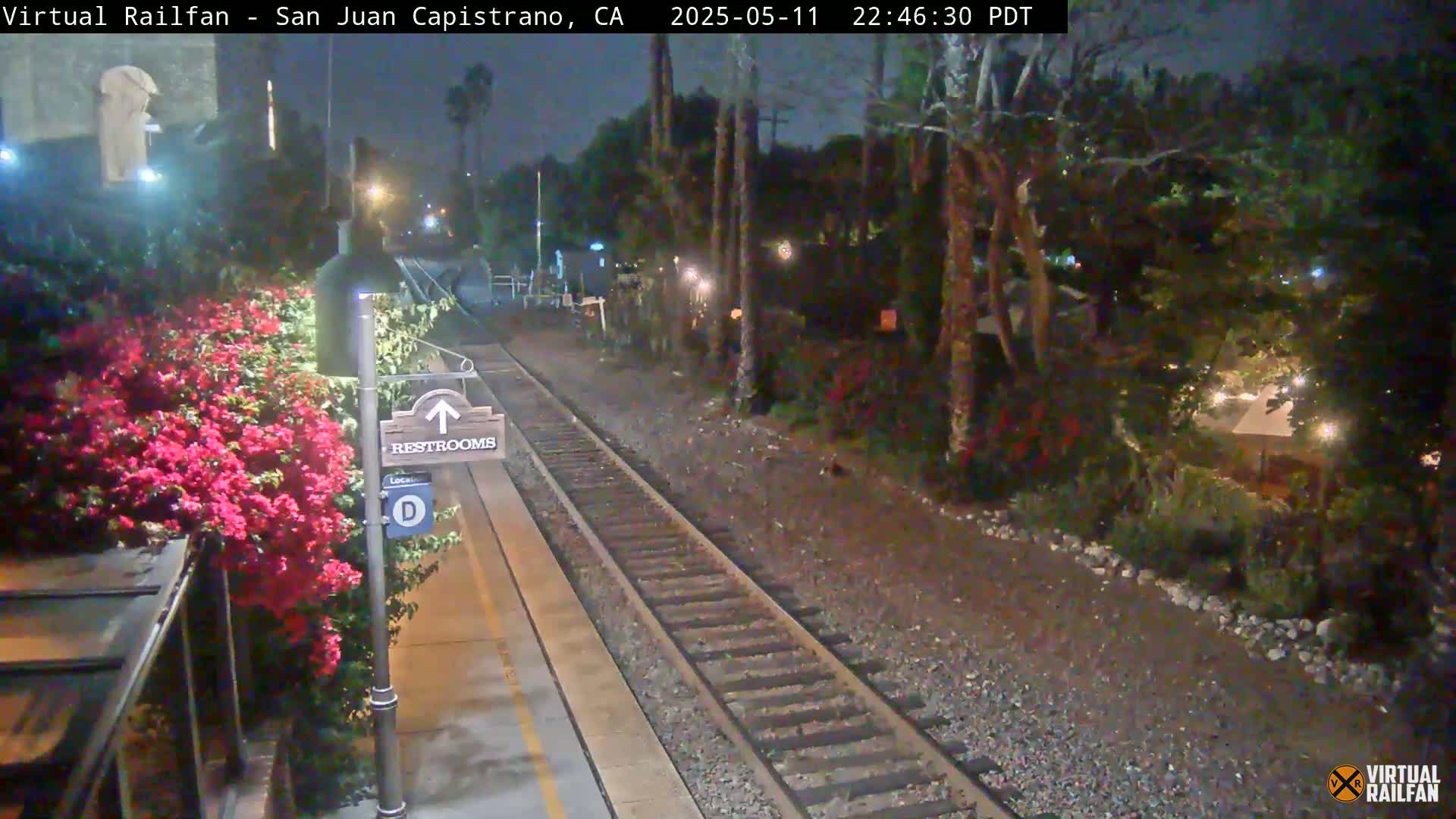 A nighttime view of a train platform with a restroom sign, vibrant pink flowers, and train tracks extending into a dimly lit landscape.