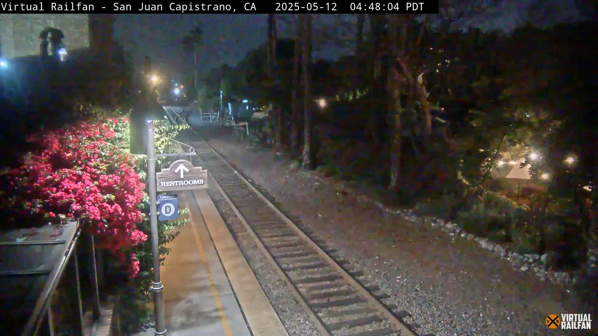 A nighttime view of a train platform with a restroom sign, blooming pink flowers, and train tracks extending into a dark, wooded area.