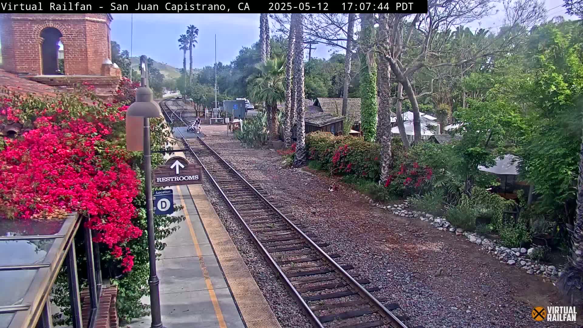 A train station platform with vibrant pink flowers, train tracks, and a partly sunny outdoor setting.