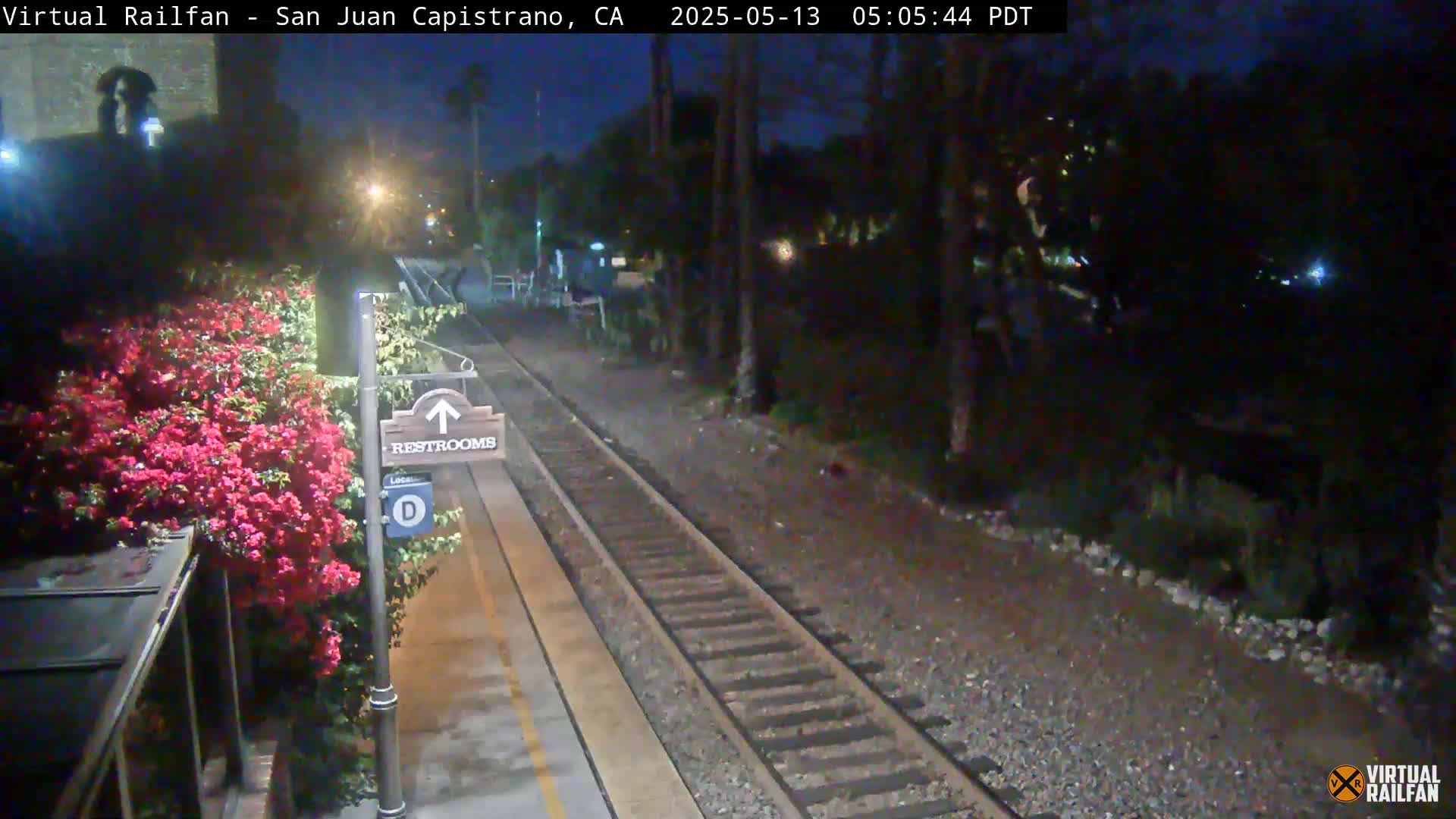 A nighttime view of a train station platform with railroad tracks, a restroom sign, and vibrant pink flowers in bloom.