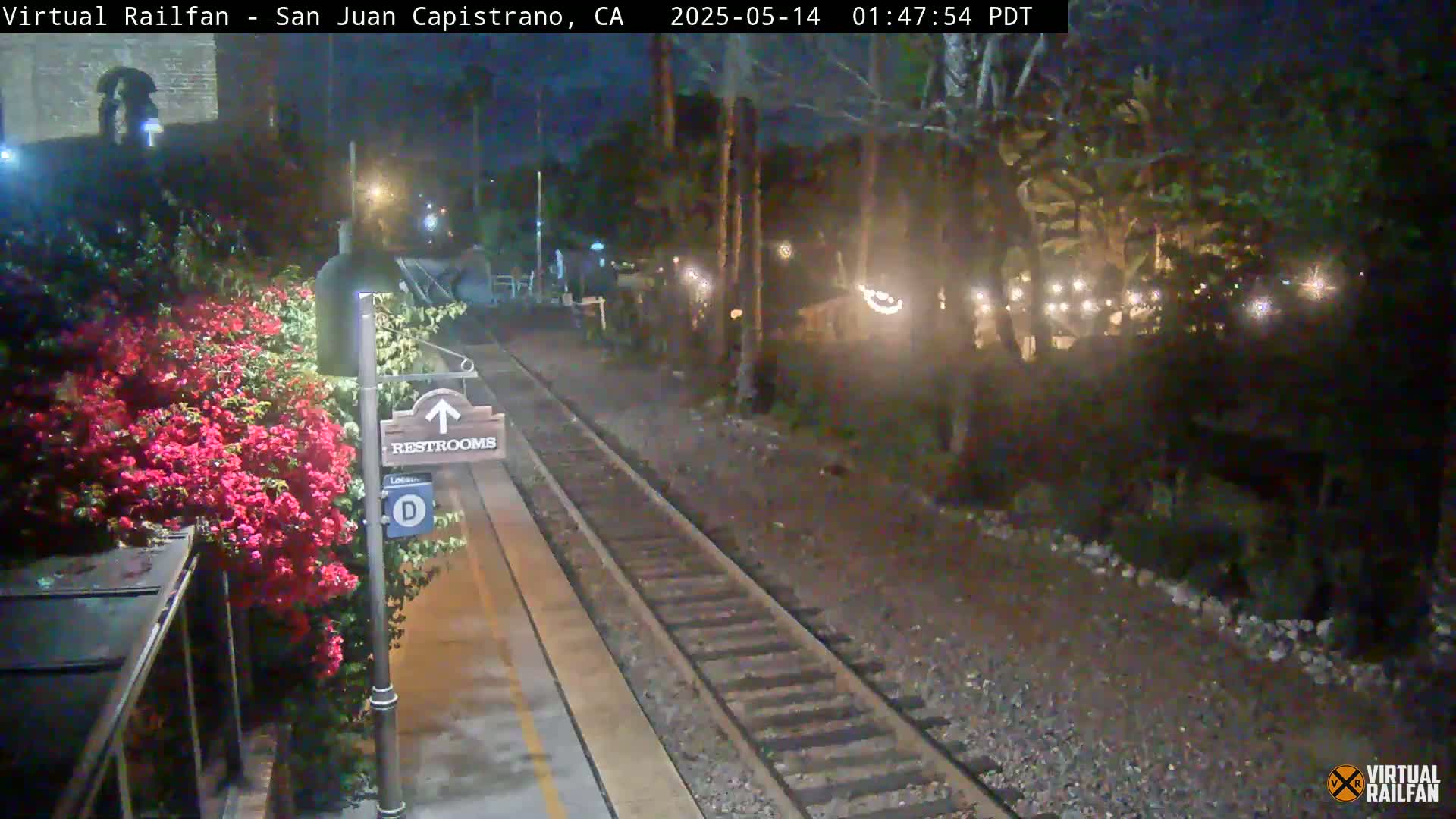 A nighttime view of a train platform with a restroom sign, blooming pink flowers, and train tracks extending into a dimly lit area with trees and string lights.