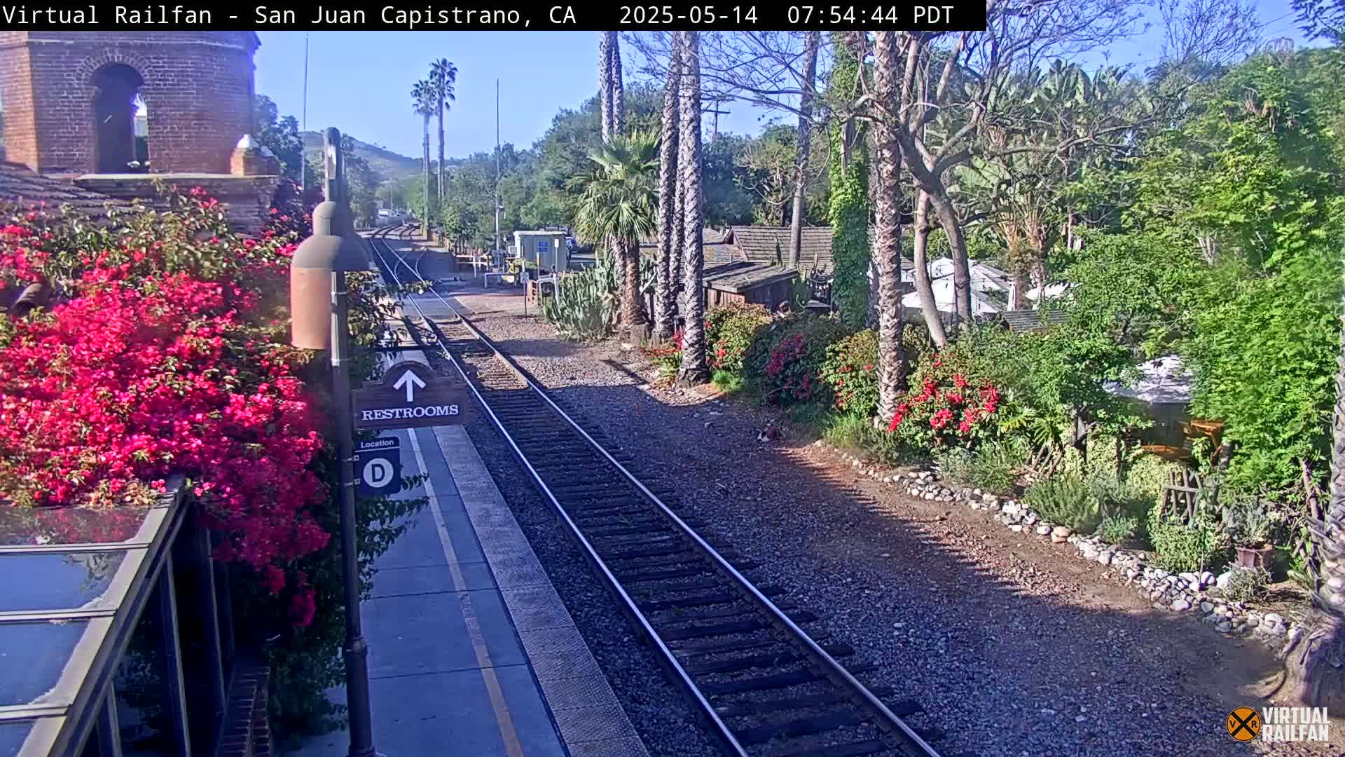 A sunny day at a train station shows a view of train tracks, lush greenery, flowering bushes, and a building with a bell tower.