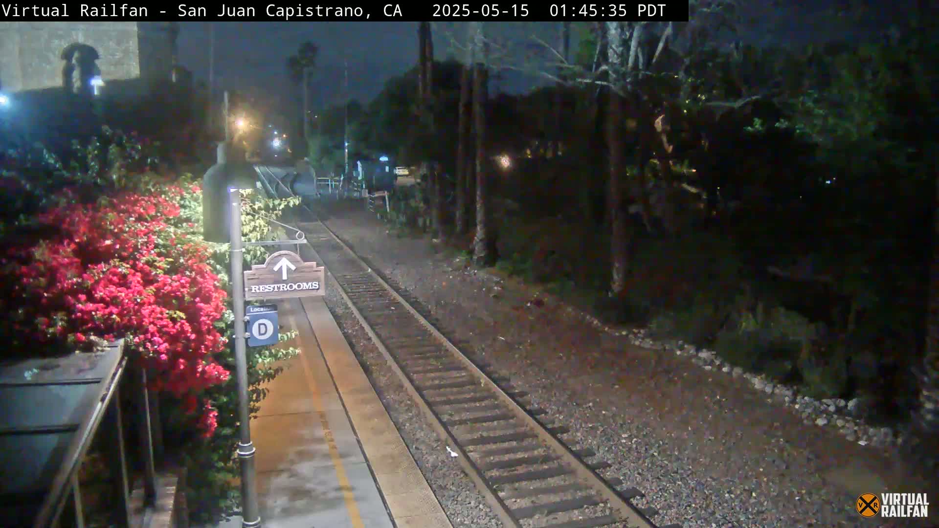 A nighttime view of a train platform with a restroom sign, vibrant red flowers, and train tracks extending into a dark, tree-lined area.