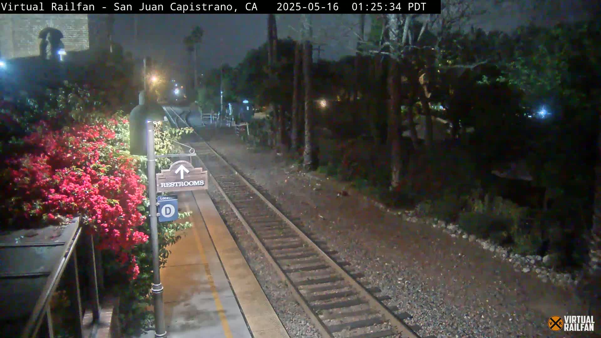 A nighttime view of a train platform with a restroom sign, vibrant red flowers, and a train track extending into a dark, tree-lined area.
