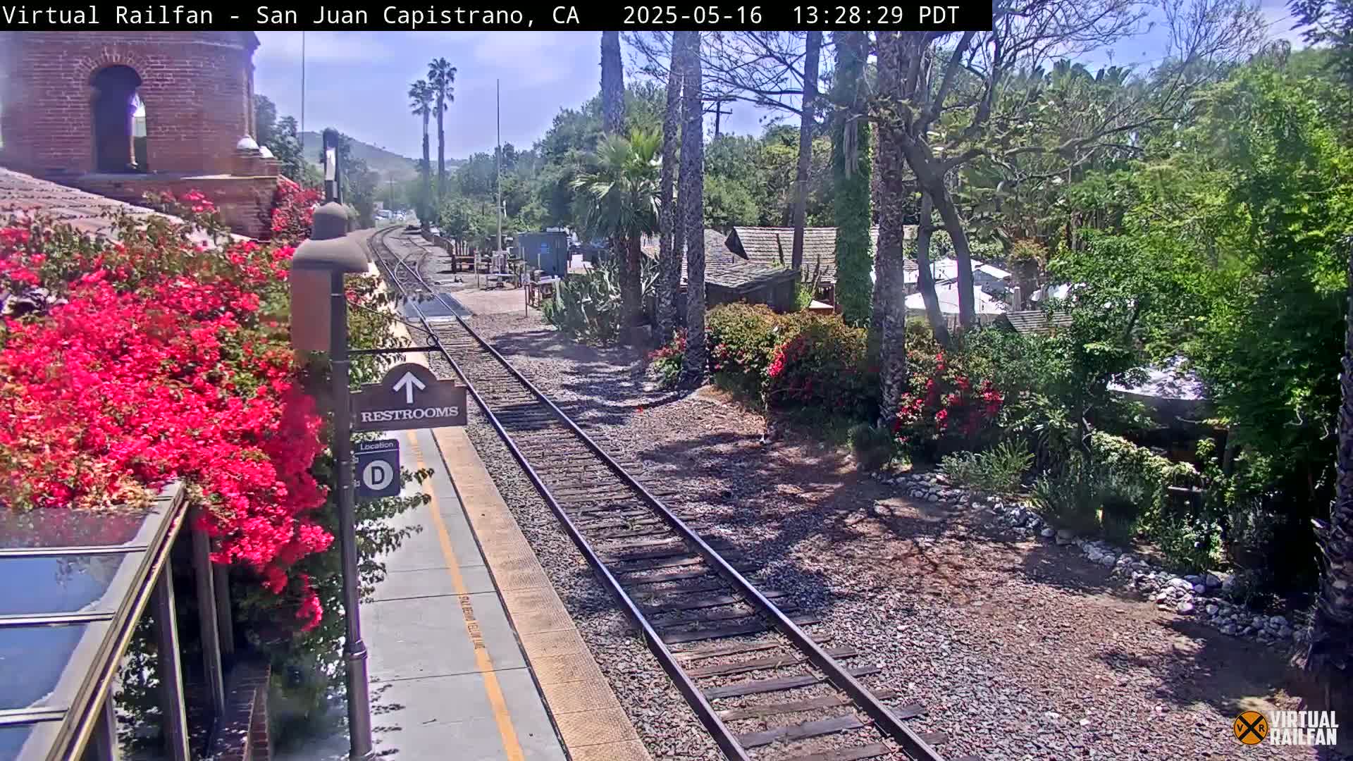A train platform with bright pink flowers is visible next to train tracks that extend into a lush, sunny landscape.