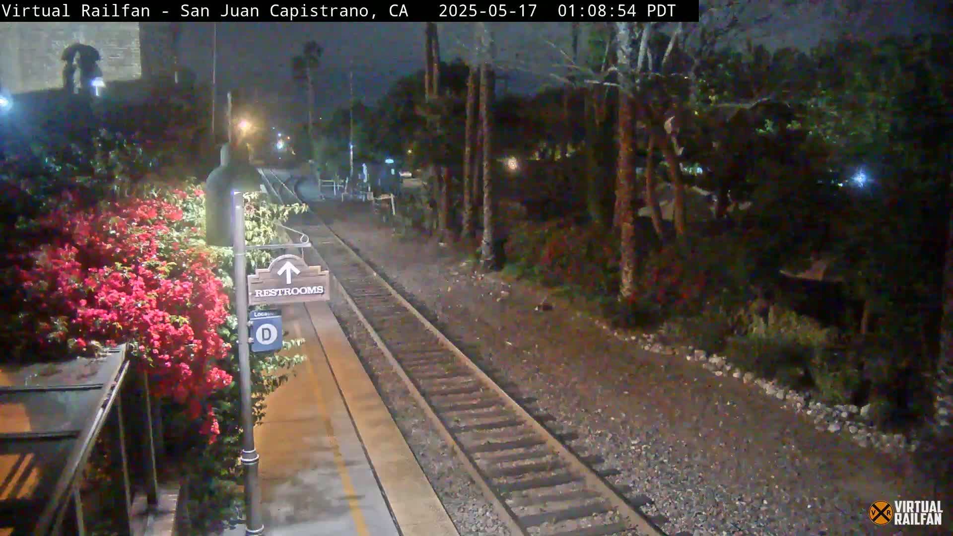 A nighttime view of a train platform with a restroom sign, blooming red flowers, and train tracks extending into a dark, tree-lined area.