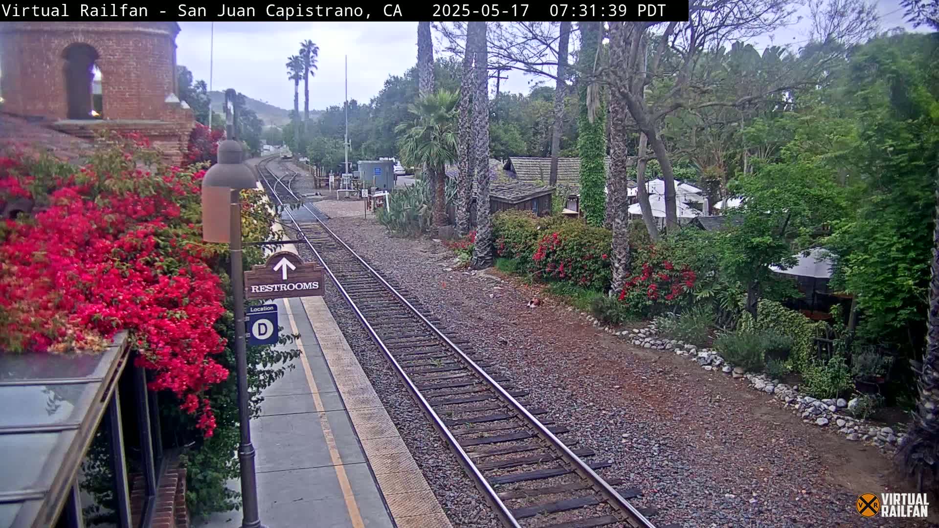 A train station platform with a restroom sign, vibrant bougainvillea, and railroad tracks, under an overcast sky.