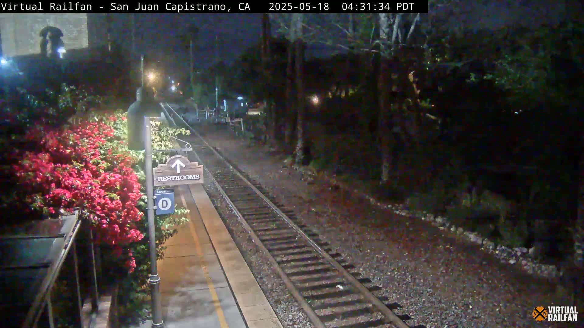 A nighttime view of a train station platform with a restroom sign, train tracks, and red flowering bushes.