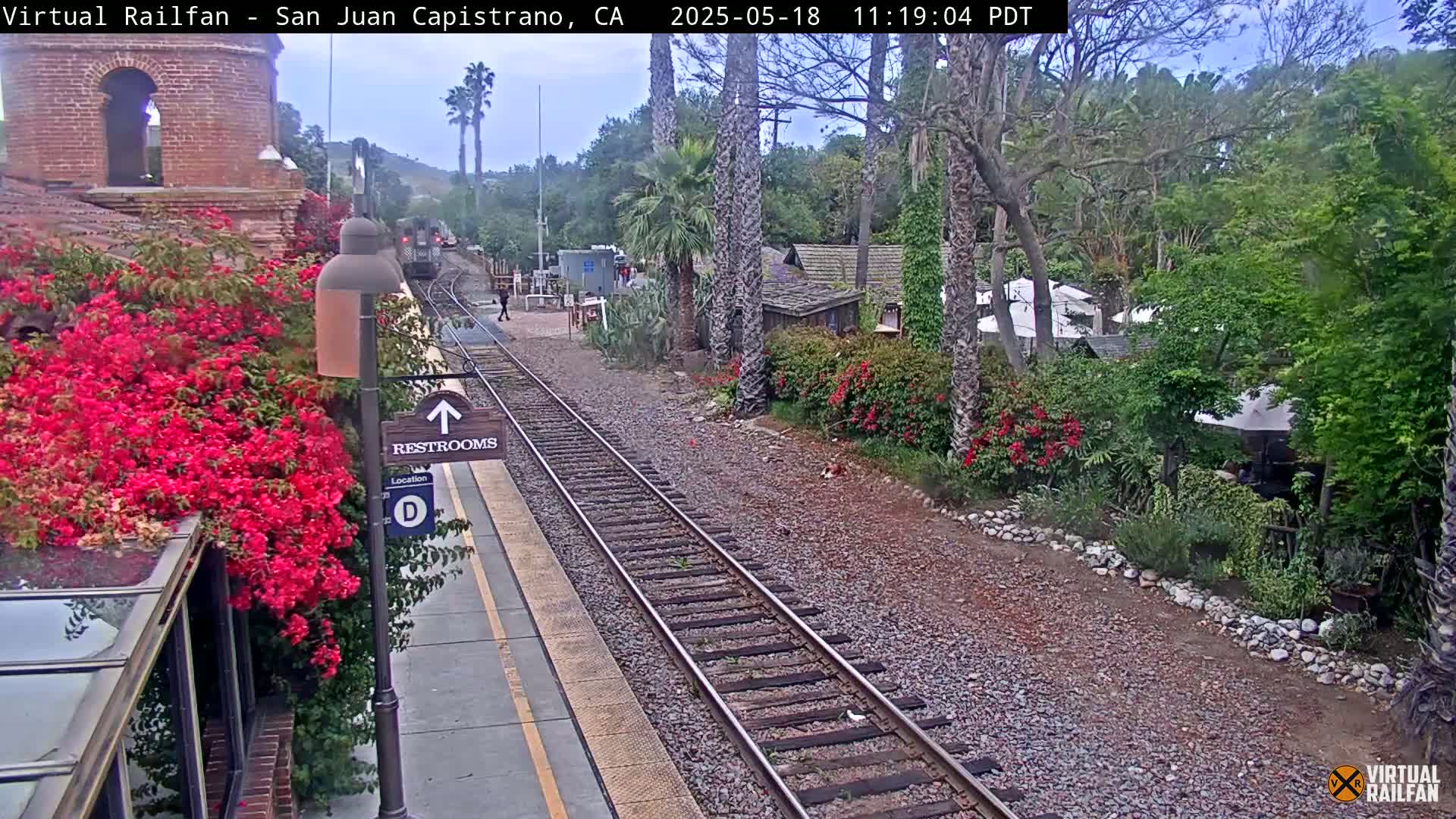 A train station platform with a train in the distance is shown, surrounded by lush greenery under an overcast sky.