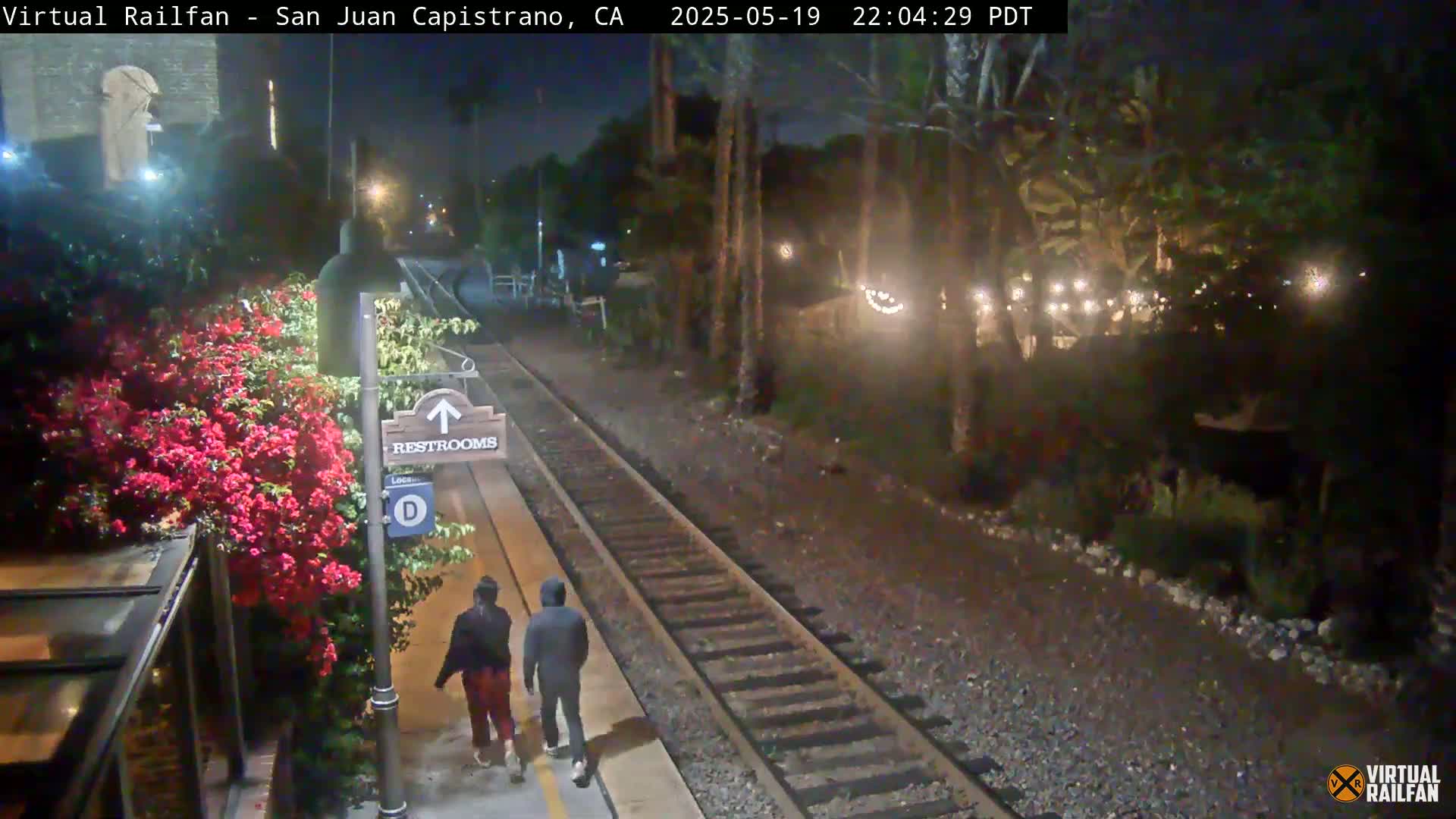 Two people walk on a train platform at night near a restroom sign and blooming red flowers, with train tracks and nighttime lights visible in the background.