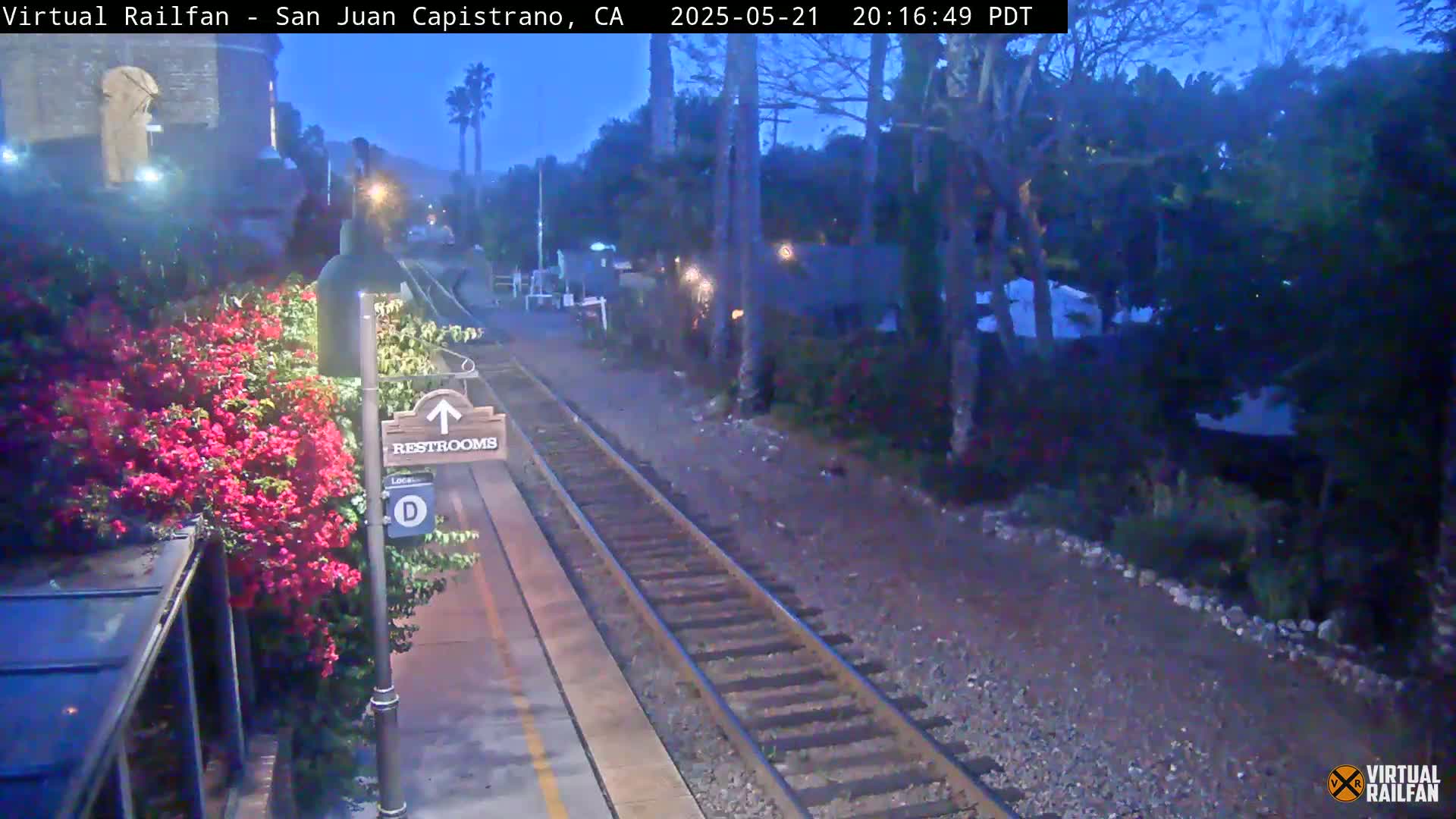 A train track at dusk runs beside a platform with vibrant pink flowers and a restrooms sign, adjacent to a building partially visible.