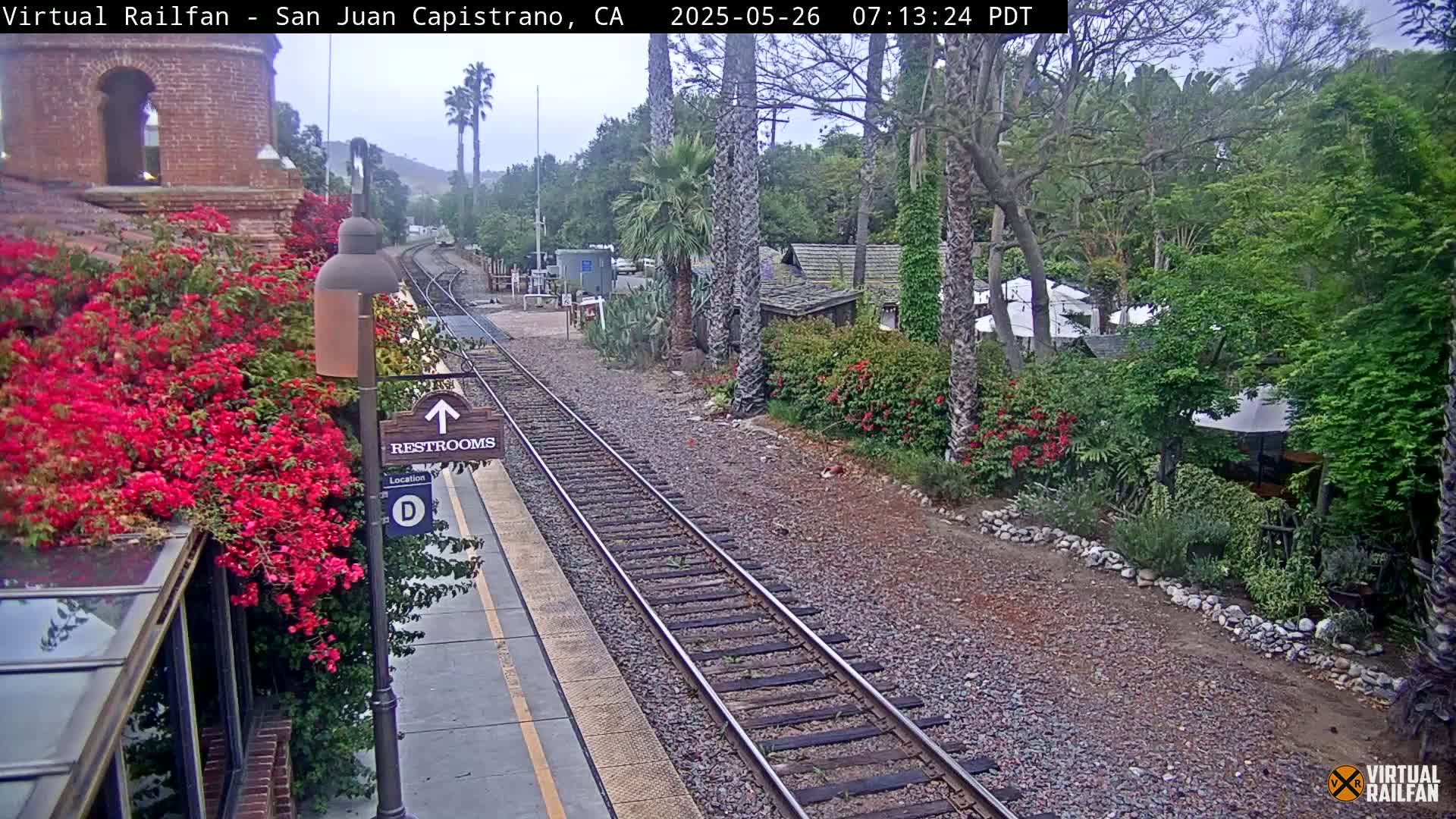 A train track runs through a lush, green landscape alongside a station platform with restrooms, under an overcast sky.