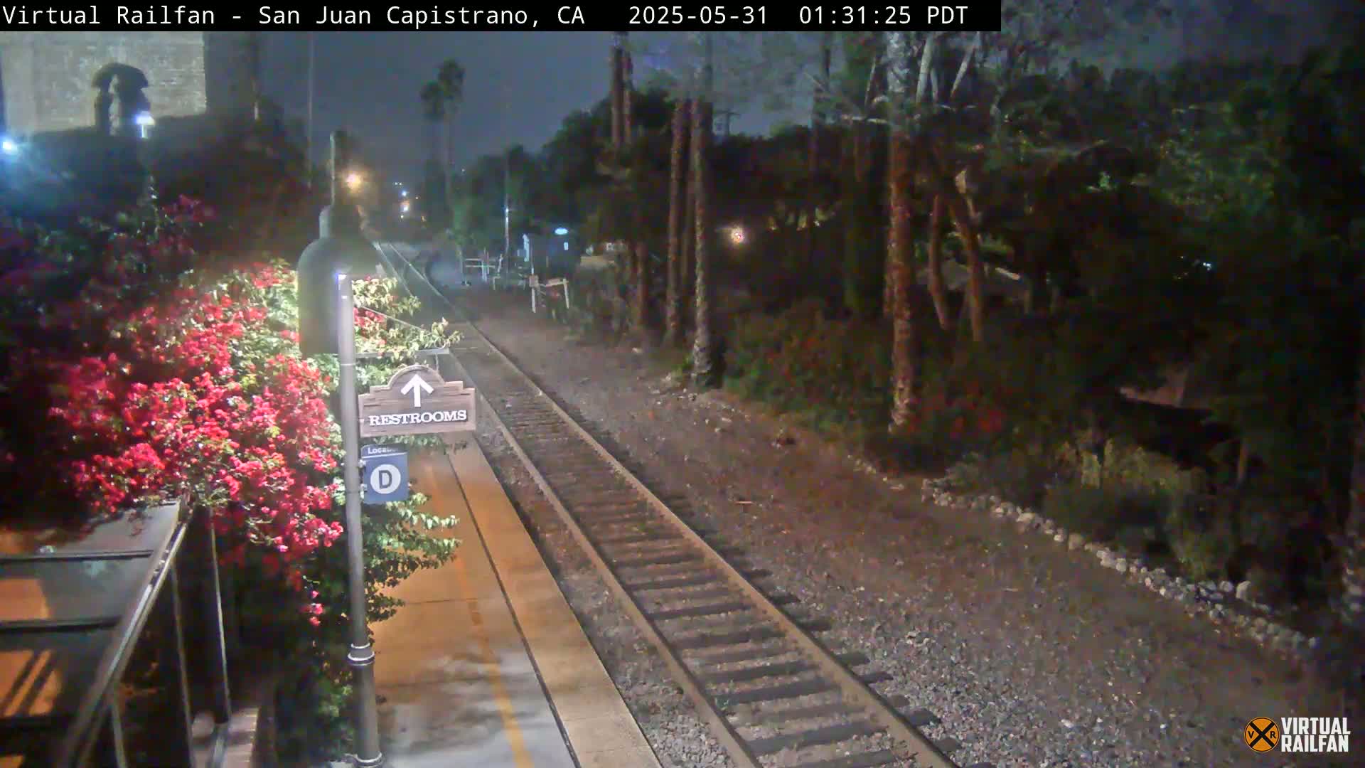 A nighttime view of a train station platform with railroad tracks, a restroom sign, and blooming red flowers next to a shelter.