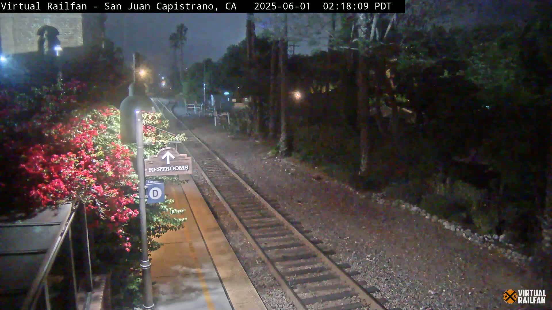 A nighttime view of a train platform with a restroom sign, blooming red flowers, and train tracks extending into a dark, tree-lined area.
