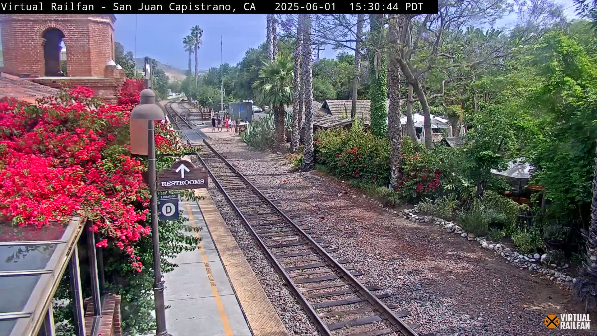 A train station platform with train tracks, surrounded by lush greenery including vibrant bougainvillea, under a sunny sky.