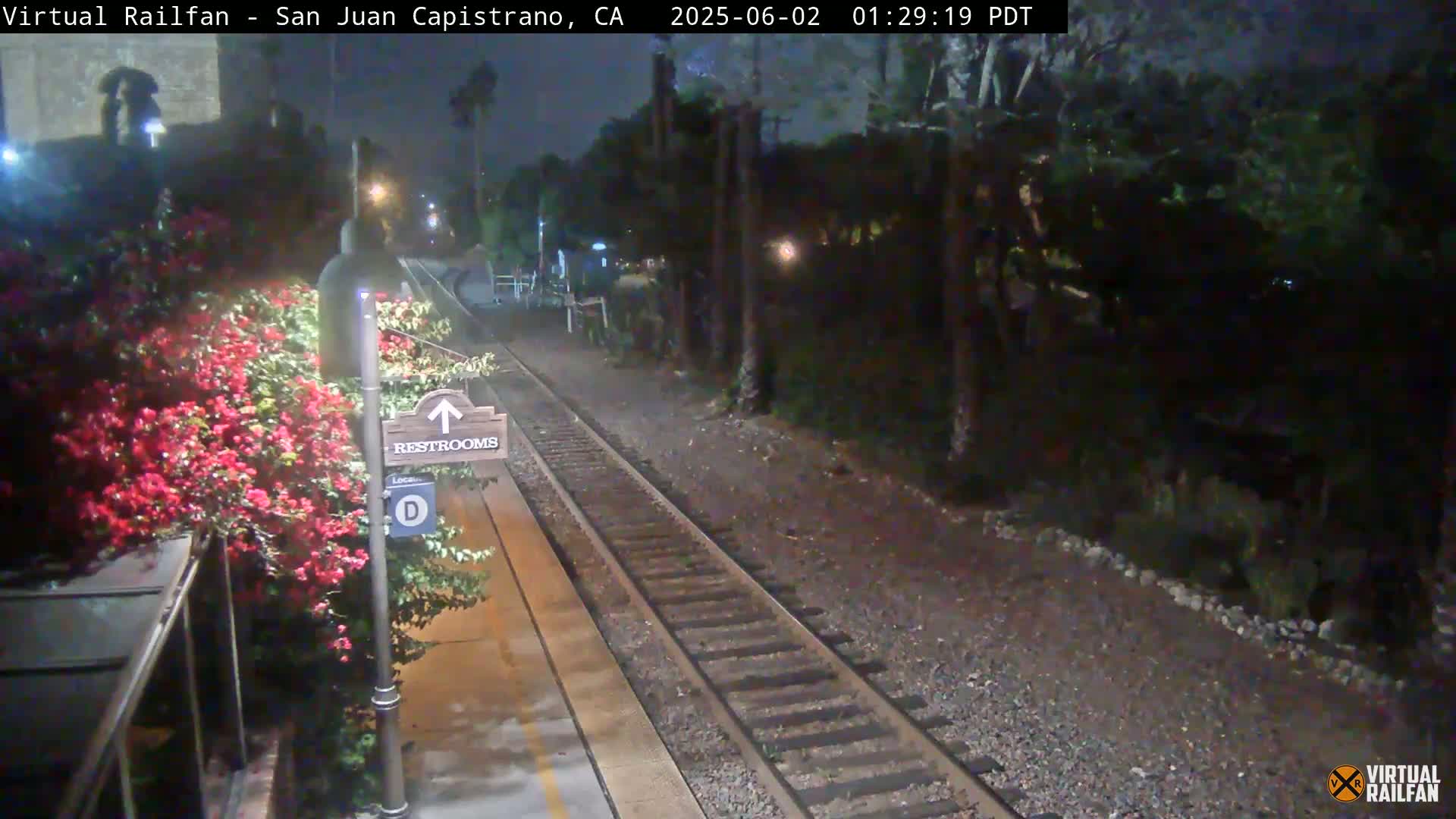 A nighttime view of a train track at a station with a restroom sign and blooming red flowers beside the platform.