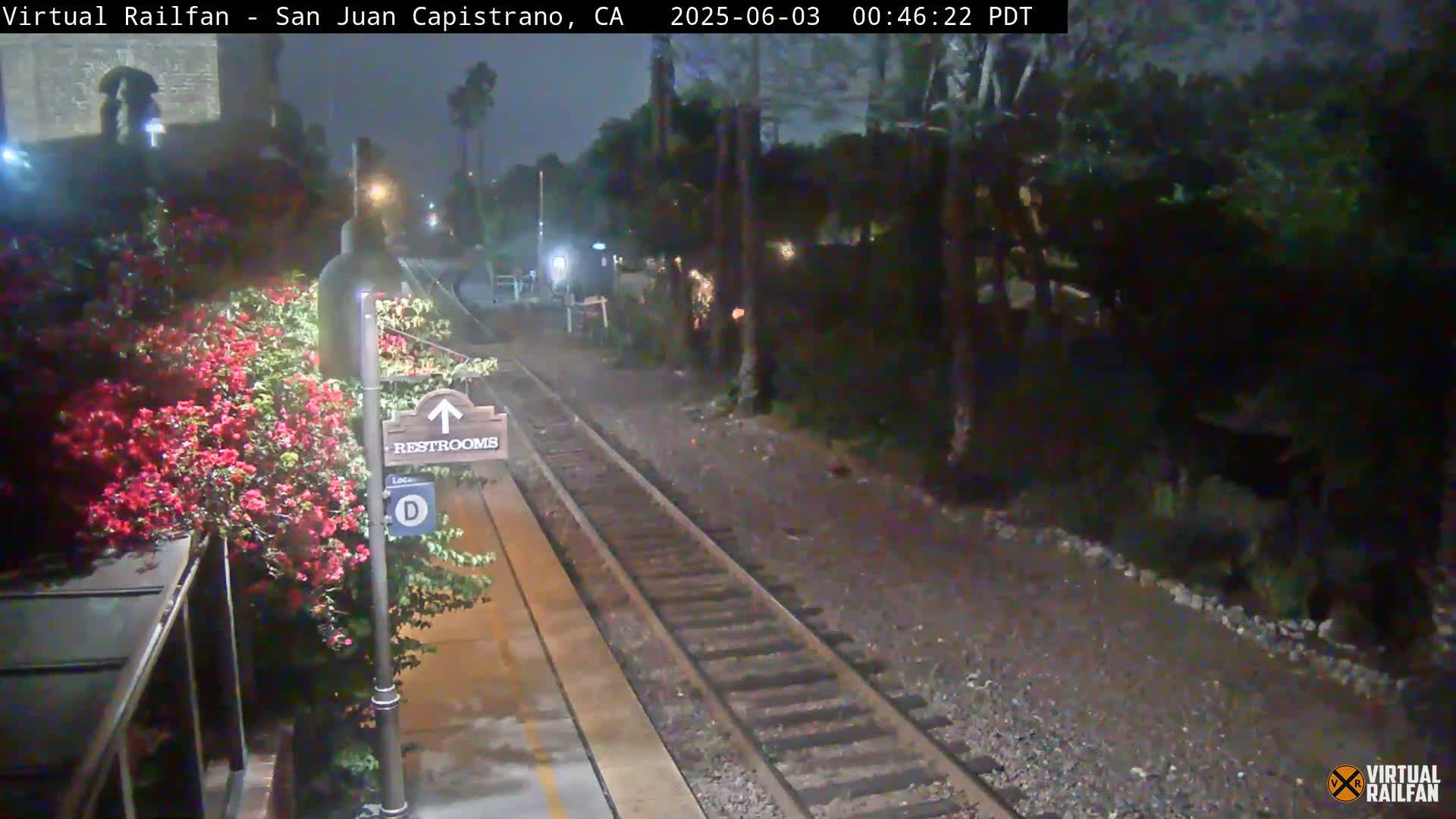 A nighttime view of a train track next to a platform with red flowers and a restrooms sign.