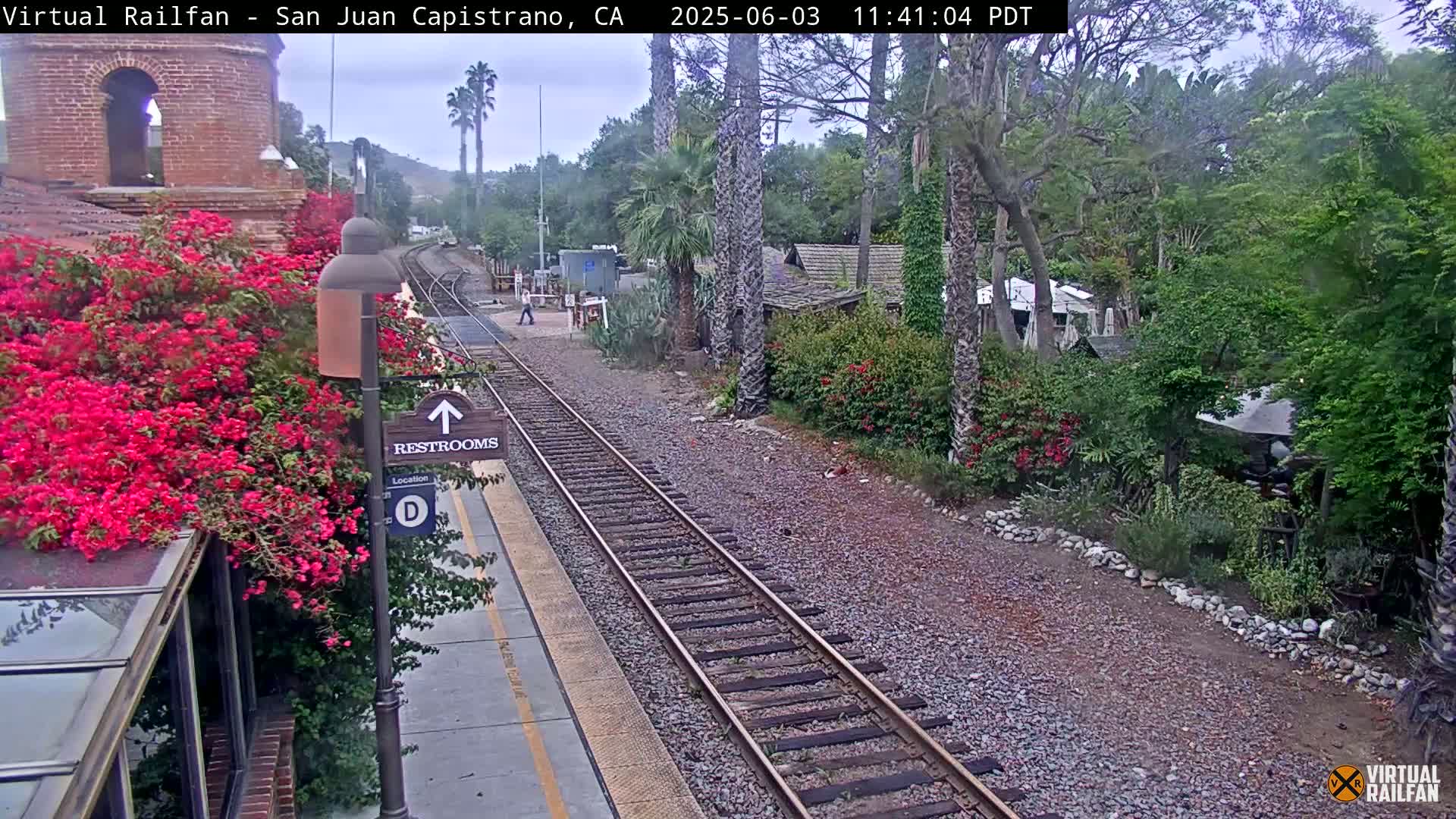 A train platform with railroad tracks, lush vegetation including vibrant bougainvillea, and a partly cloudy sky.