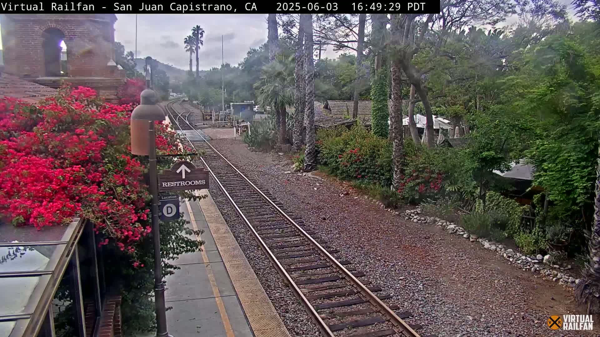 A train station platform with vibrant red flowers, a restroom sign, and train tracks under an overcast sky.
