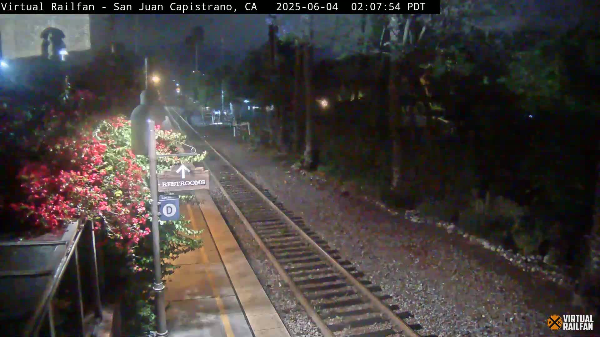 A nighttime view of a train station platform with railroad tracks, a restroom sign, and flowering bushes.