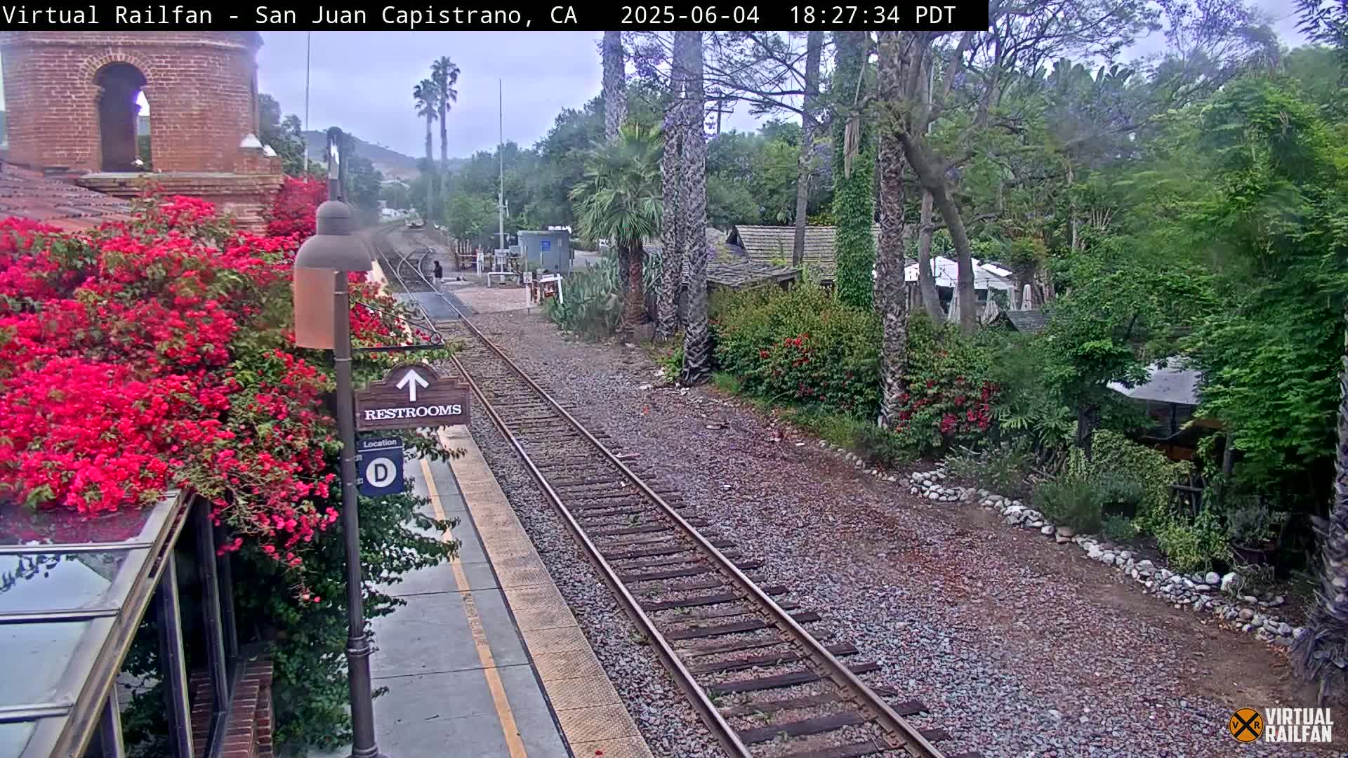 A train track runs through a landscaped area with lush vegetation, including vibrant red flowers and palm trees, under an overcast sky.