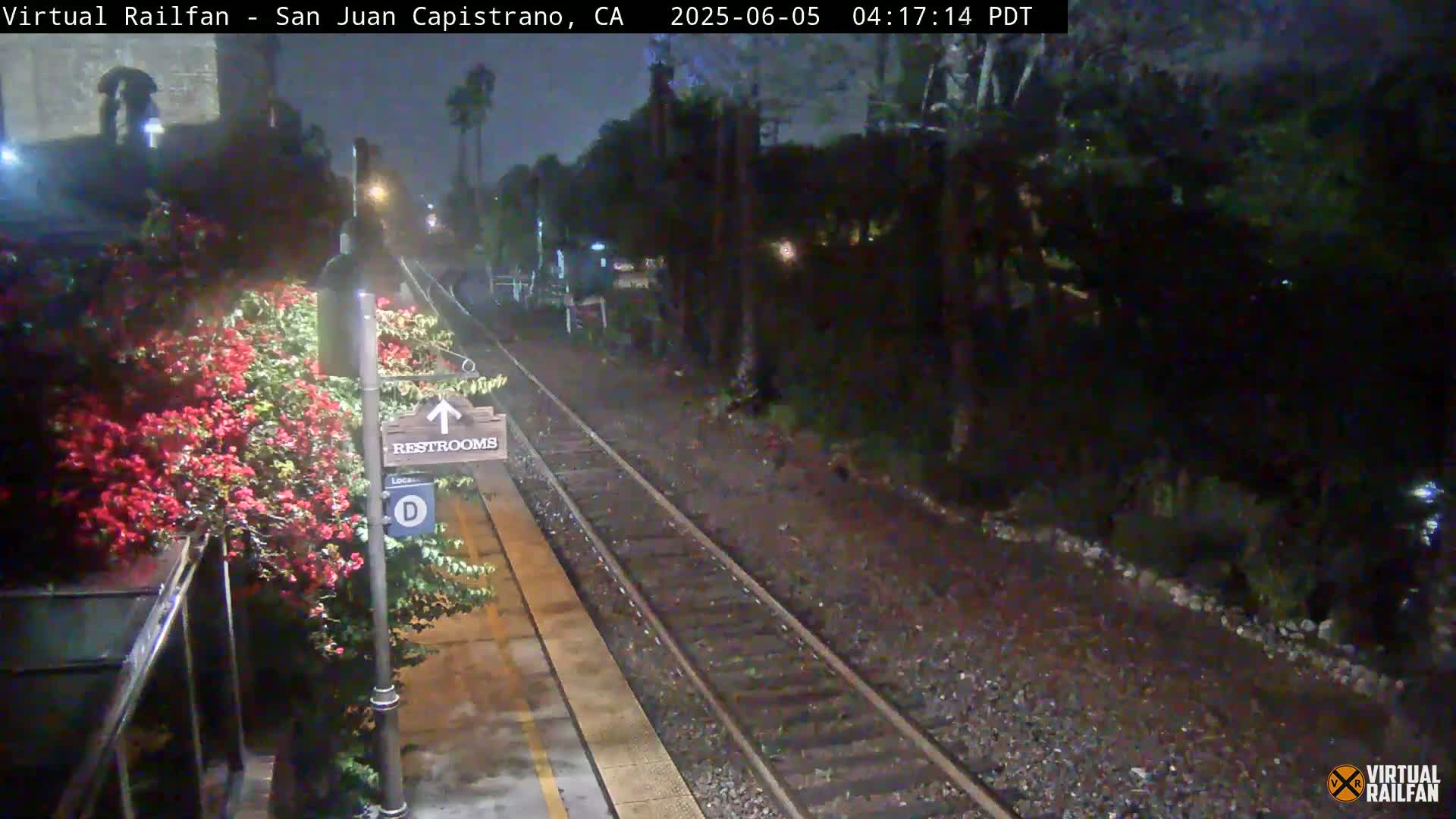 A nighttime view of a train platform with a restroom sign, lush flowering bushes, and train tracks extending into a dark, tree-lined area under a misty sky.
