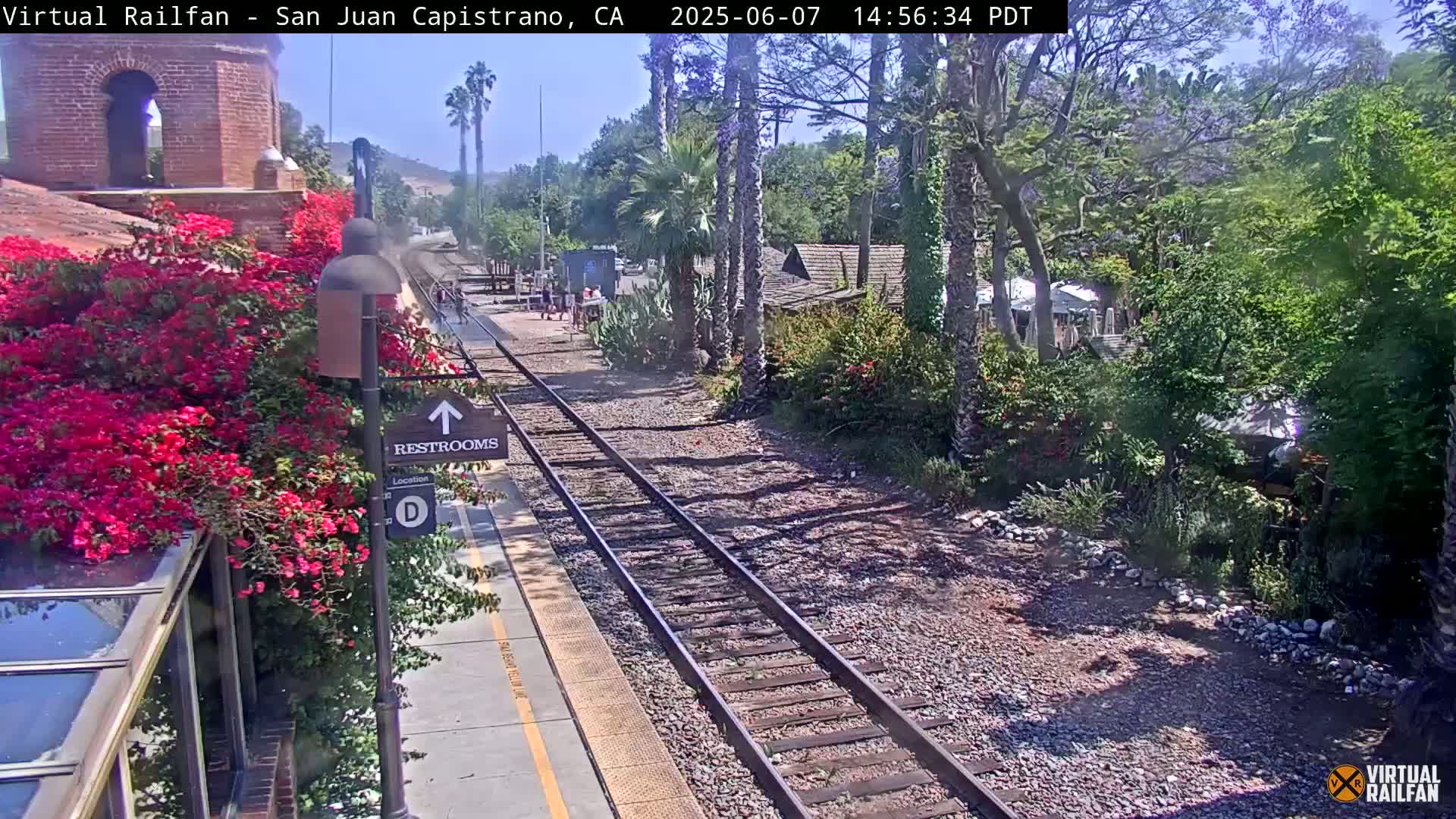 A sunny train platform with a vibrant bougainvillea bush is next to a set of train tracks that lead through lush greenery and small buildings.