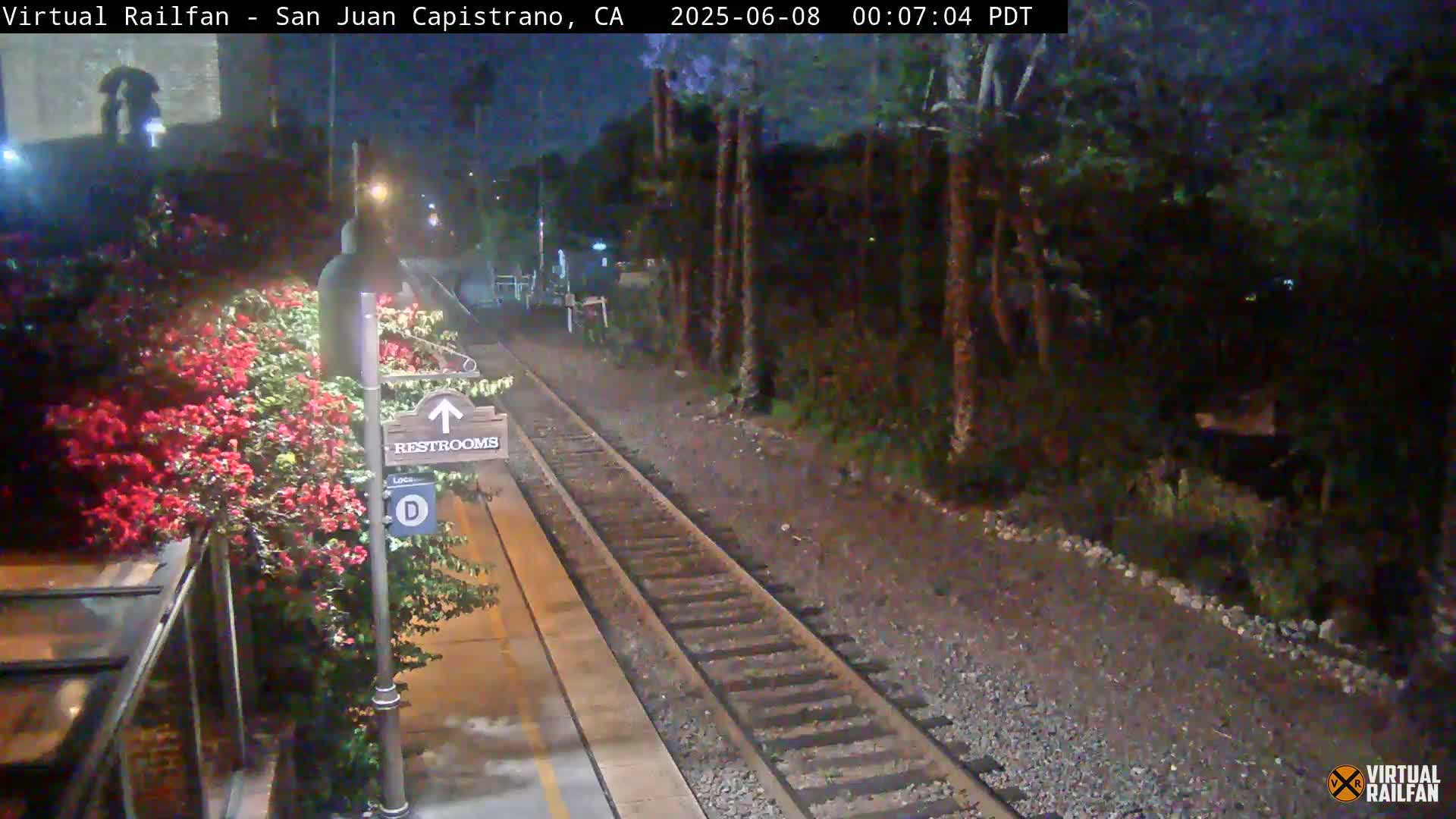 A nighttime view of a train platform with a restroom sign, blooming flowers, and train tracks extending into a dark, tree-lined area.