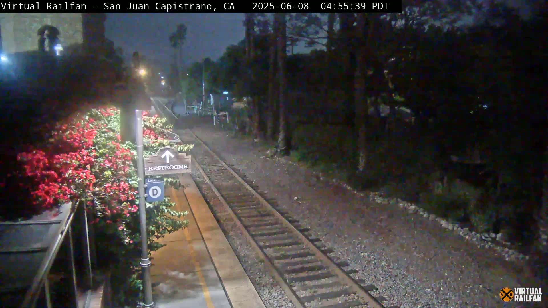 A nighttime view of a train track and platform with a restroom sign, vibrant red flowers, and dark foliage surrounding the area.