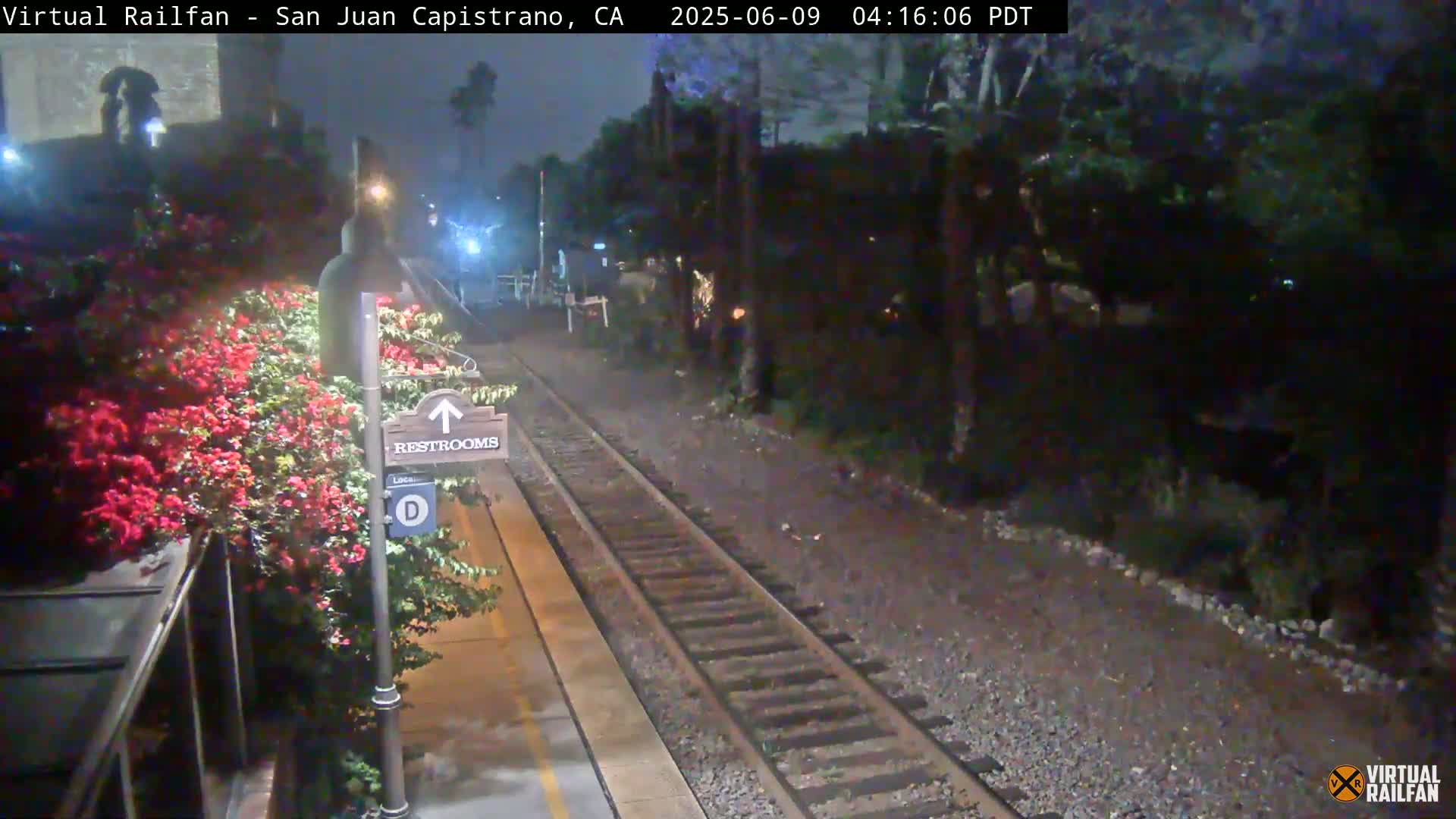 A nighttime view of a train station platform with railroad tracks, a restroom sign, and blooming red flowers next to a shelter.