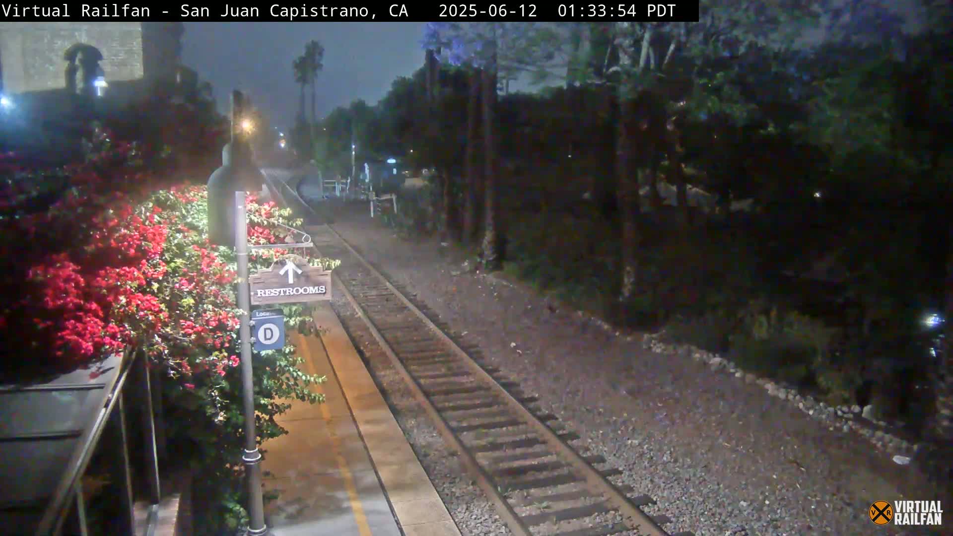 A nighttime view of a train platform with a restroom sign, vibrant red flowers, and train tracks extending into a dark, tree-lined area.