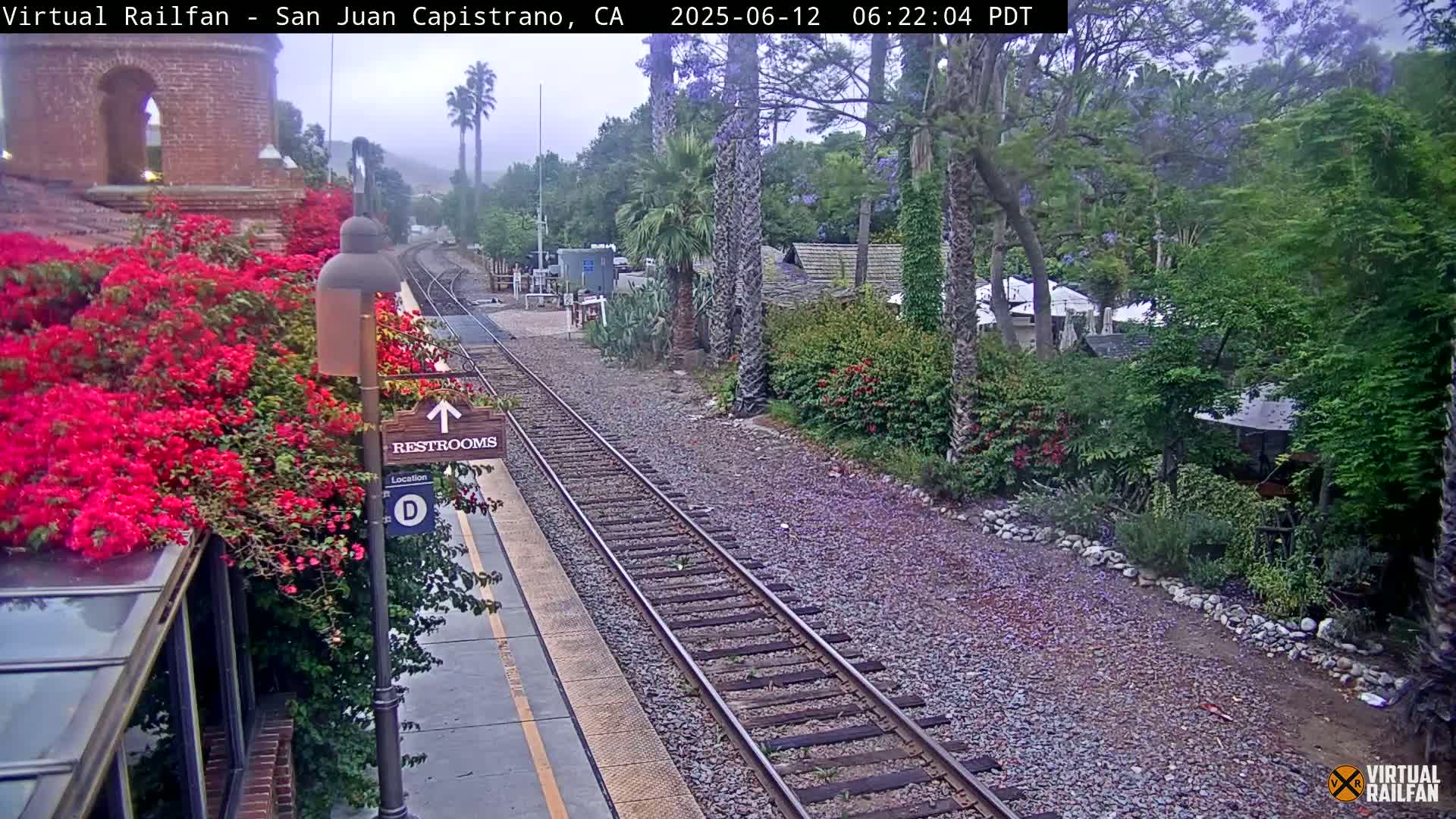 A train track runs through a station platform next to lush greenery, including vibrant red flowers, under an overcast sky.