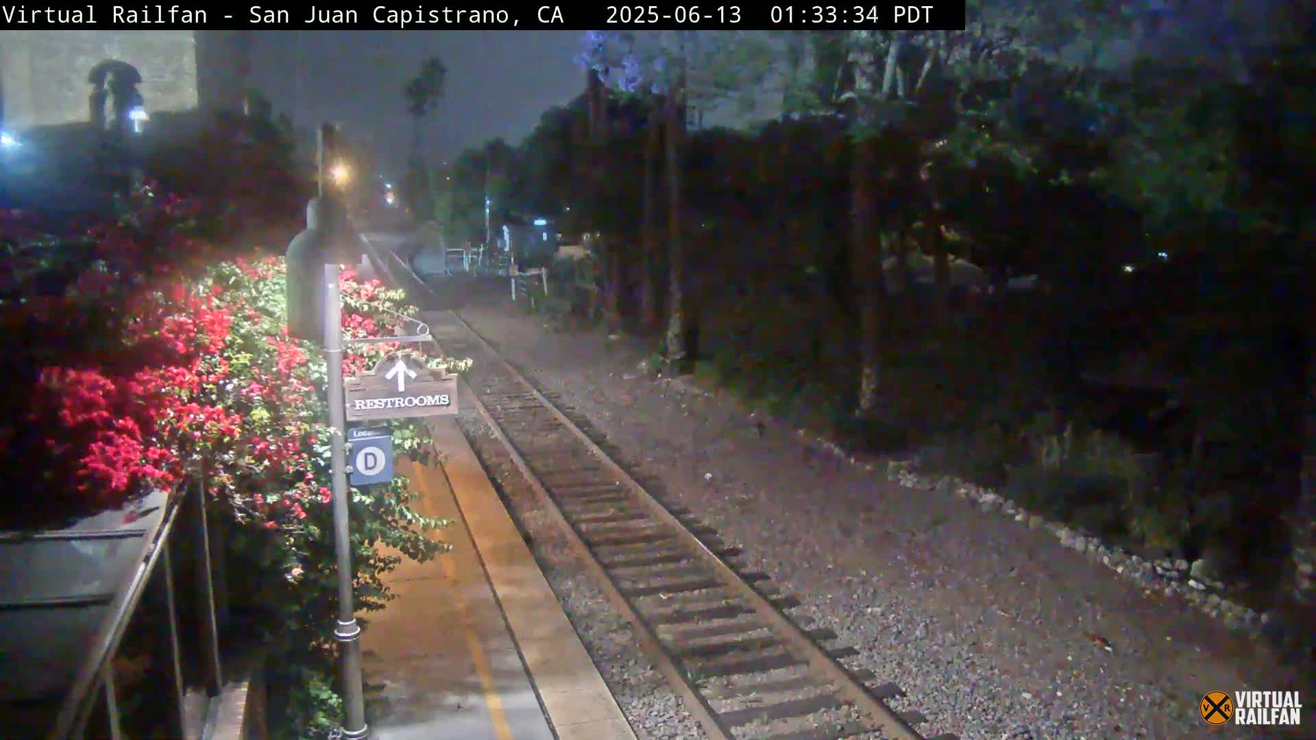 A nighttime view of a train station platform with a restroom sign, vibrant red flowers, and train tracks extending into a dark, tree-lined area.