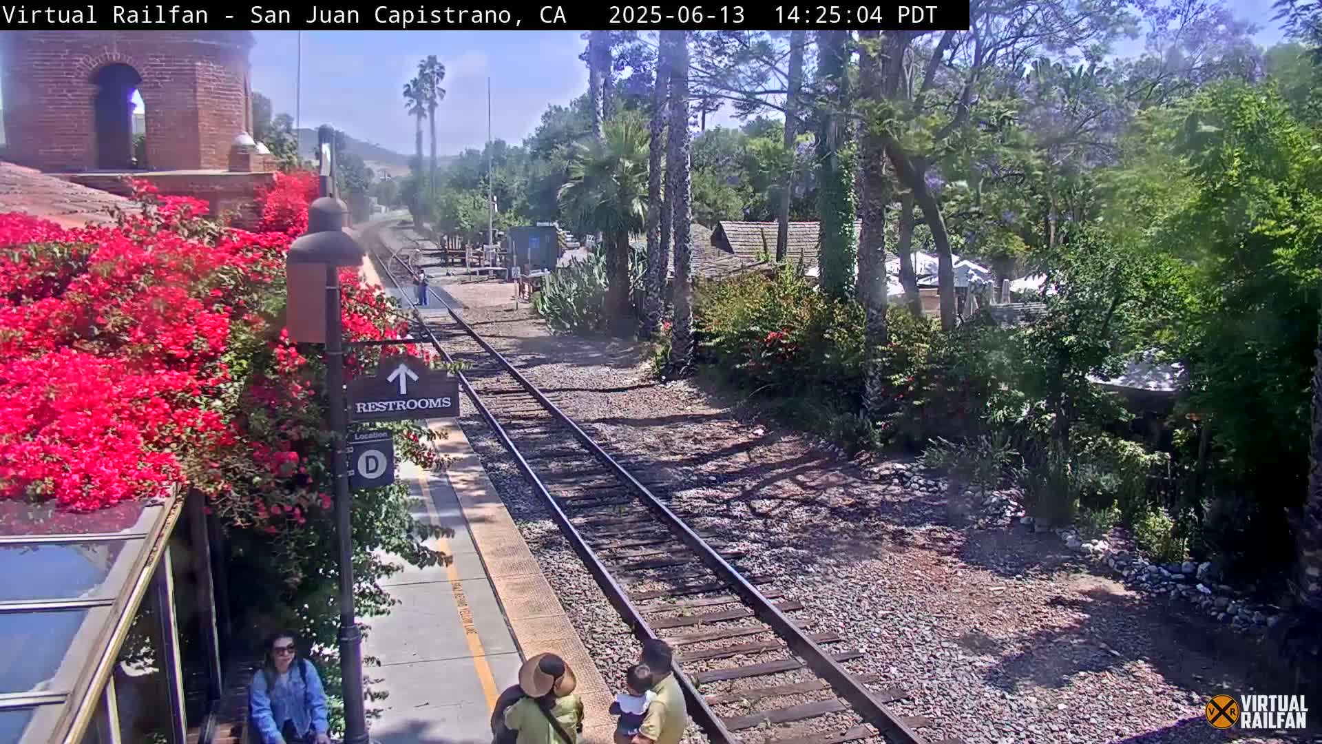 A sunny day at a train station shows a railway track, a platform with several people, vibrant bougainvillea bushes, and lush greenery surrounding the area.