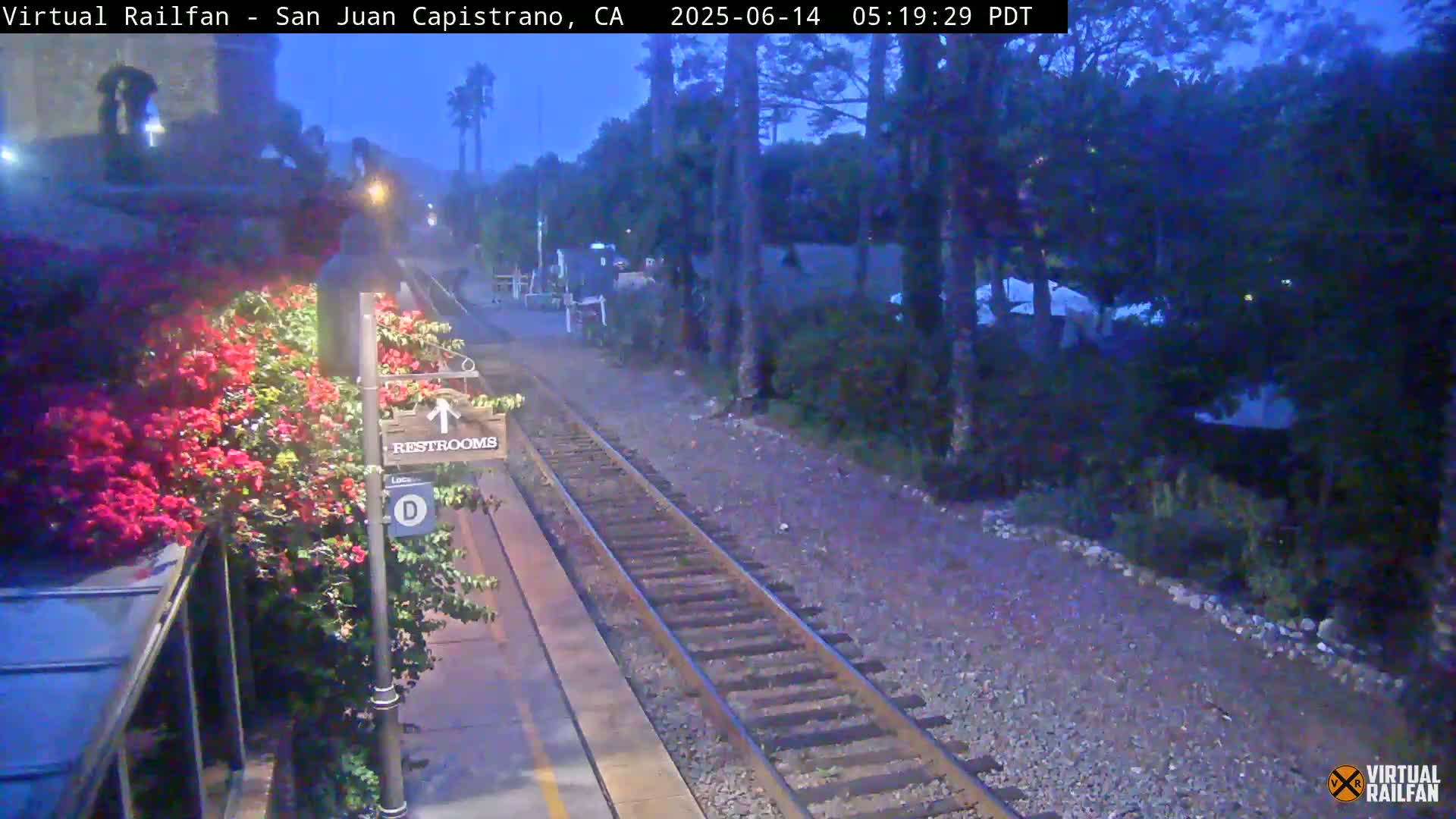 A train track at twilight runs alongside a platform with vibrant pink flowers, a restroom sign, and a few structures in the background.