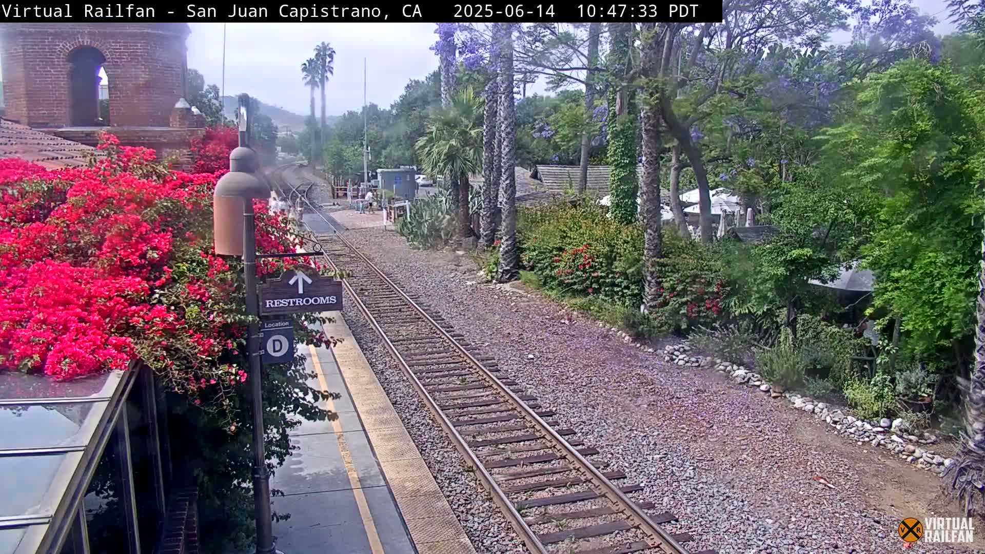 A train station platform with vibrant pink bougainvillea next to a restroom sign, railroad tracks, and lush green landscaping under a partly cloudy sky.
