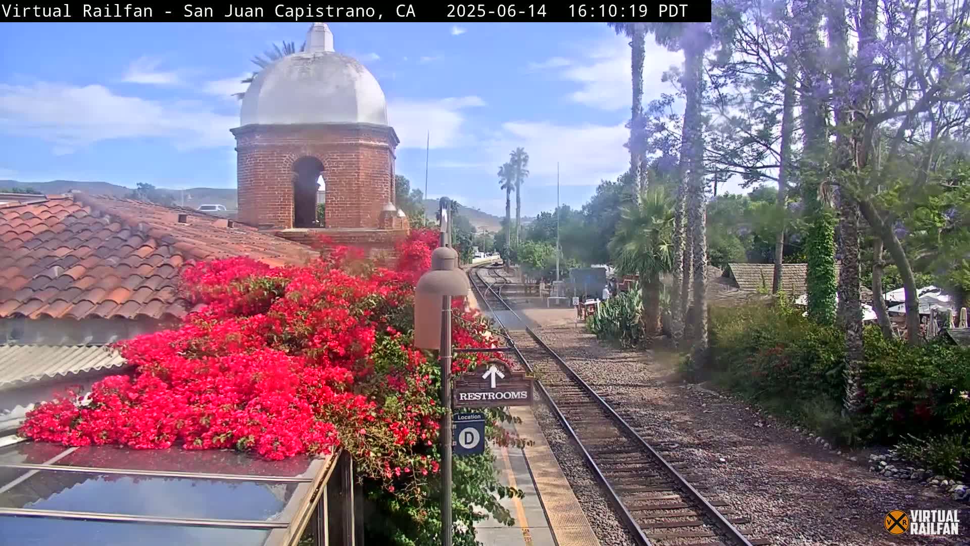 A train station platform with vibrant pink bougainvillea next to a restroom sign, railroad tracks, and lush green landscaping under a partly cloudy sky.