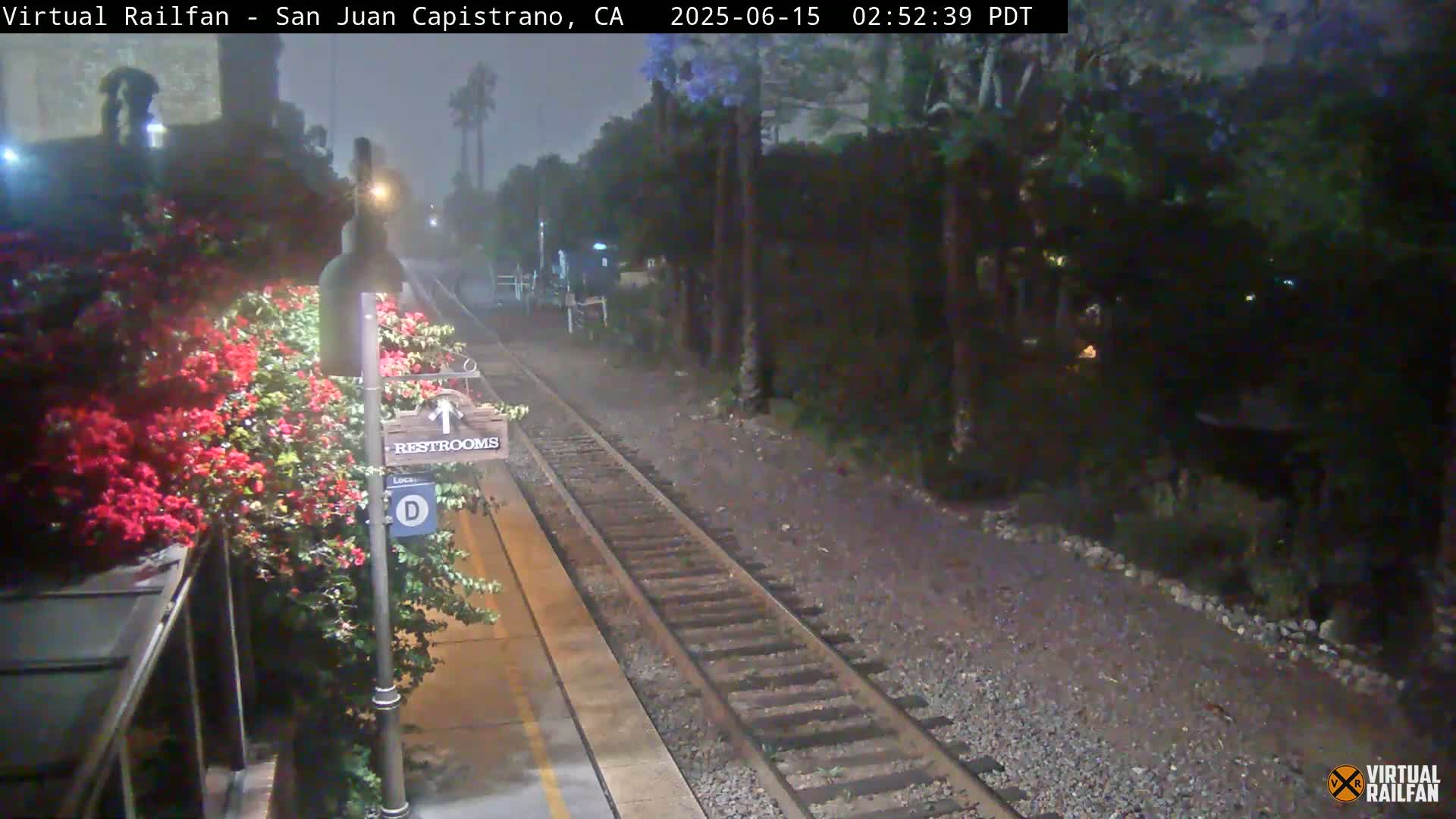 A nighttime view of a train station platform with a restroom sign, blooming red flowers, and train tracks extending into a foggy, dark background.