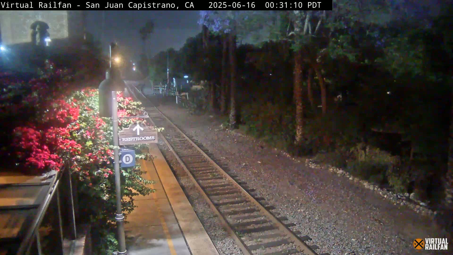 A nighttime view of a train track at a station with a restroom sign and blooming flowers next to the platform under hazy conditions.