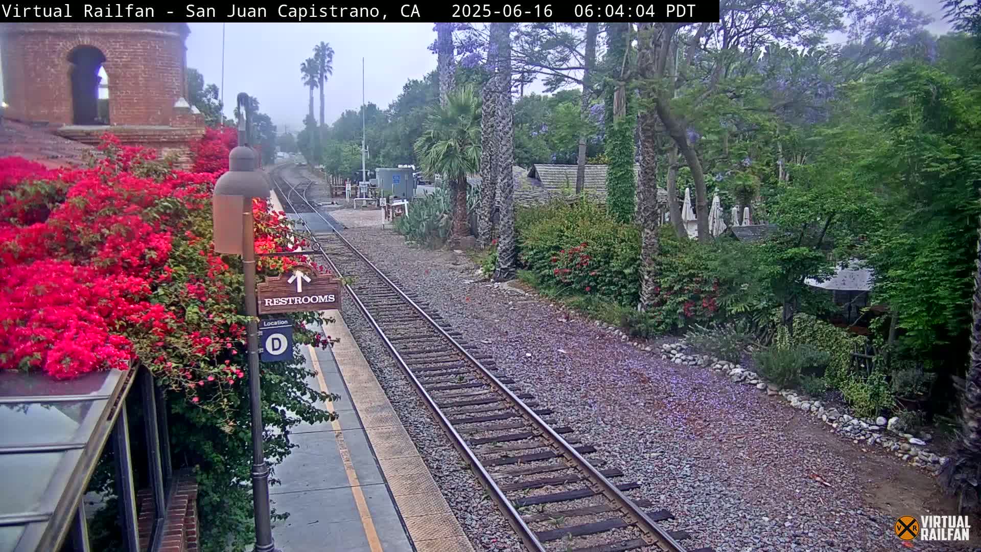 A train station platform with vibrant red flowers, a restroom sign, and train tracks, all under an overcast sky.
