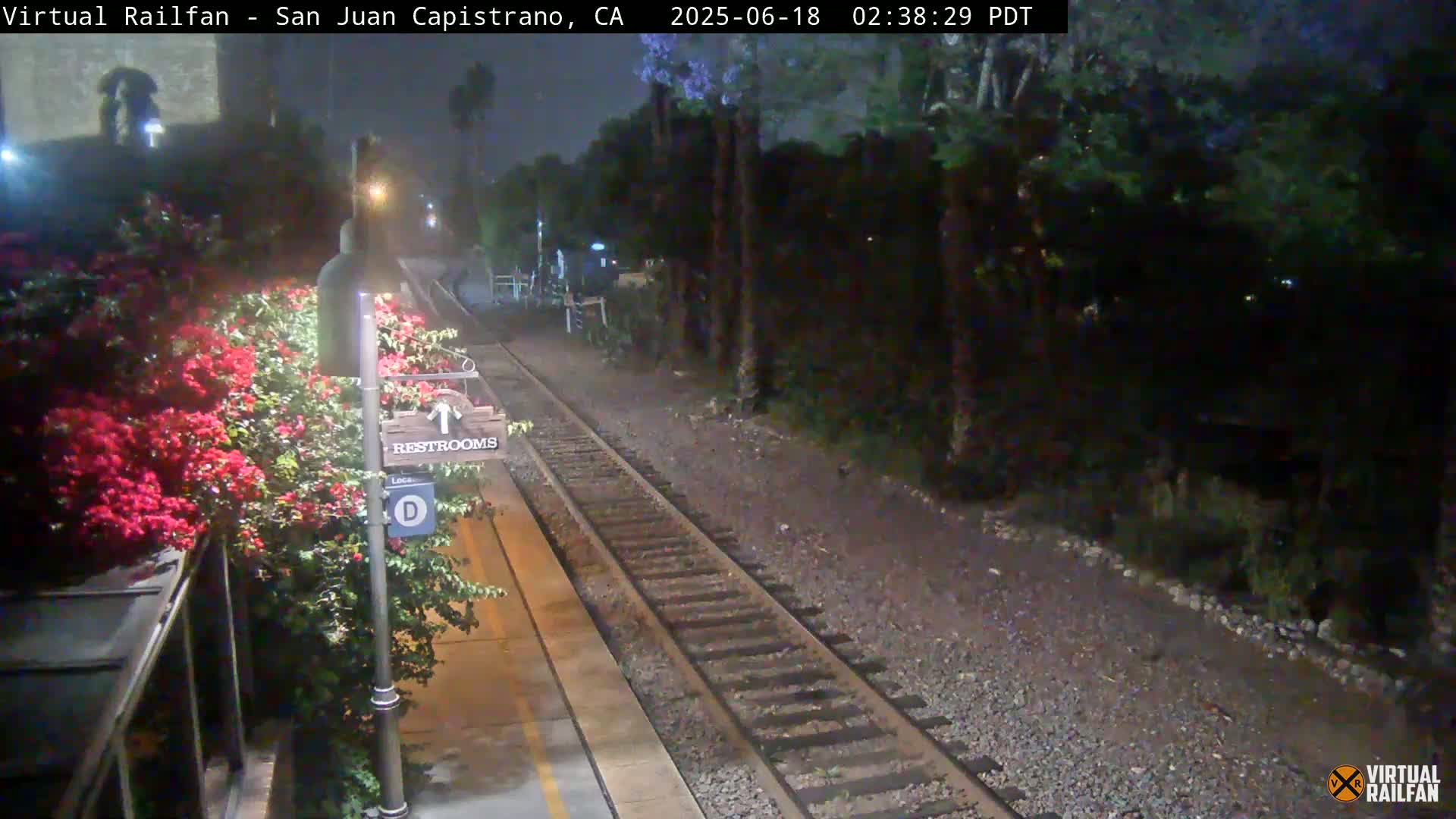 A nighttime view of a train track at a station with blooming bougainvillea next to a restroom sign.