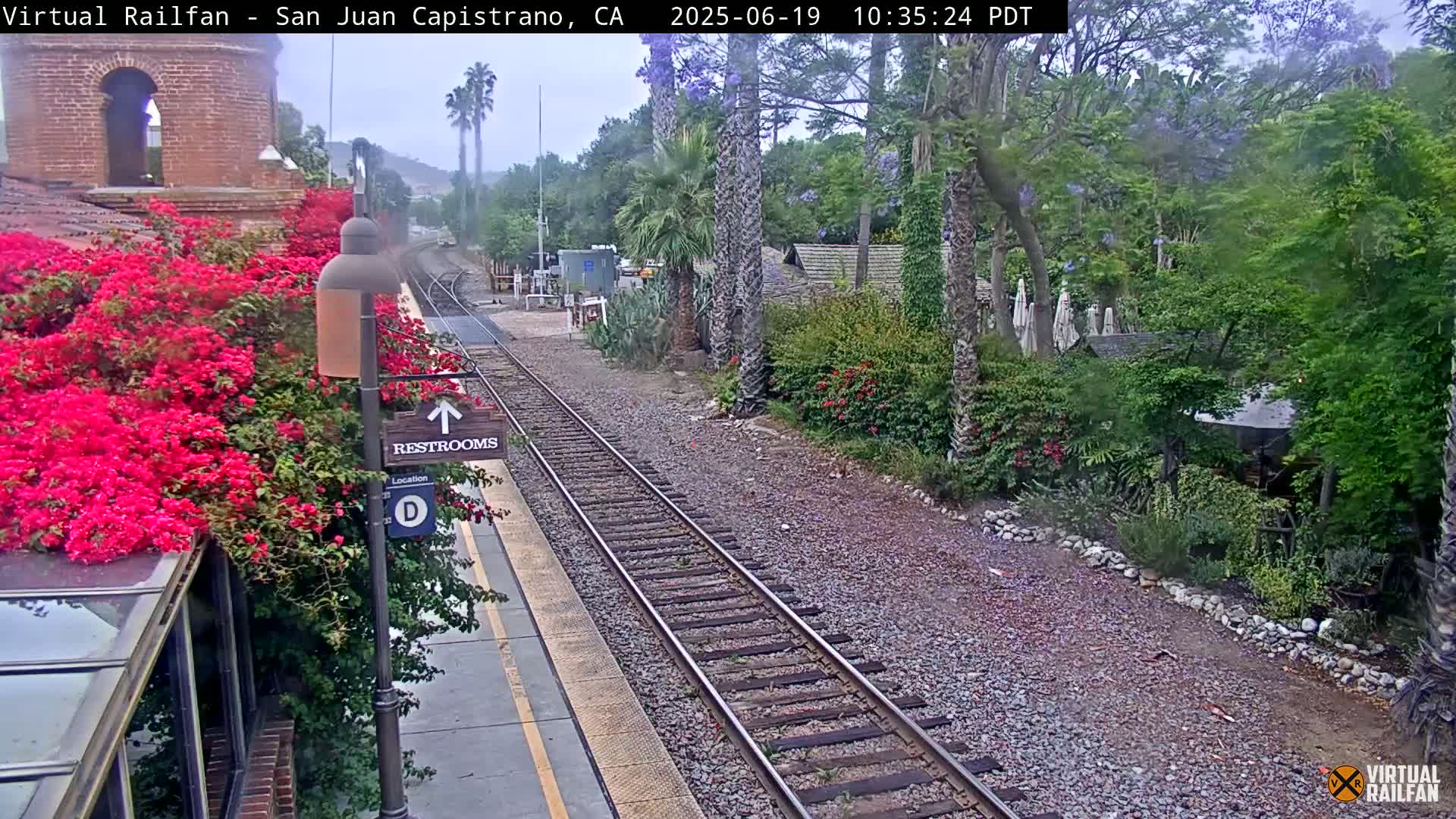 A train station platform with vibrant pink flowers, railroad tracks, and lush green landscaping under an overcast sky.