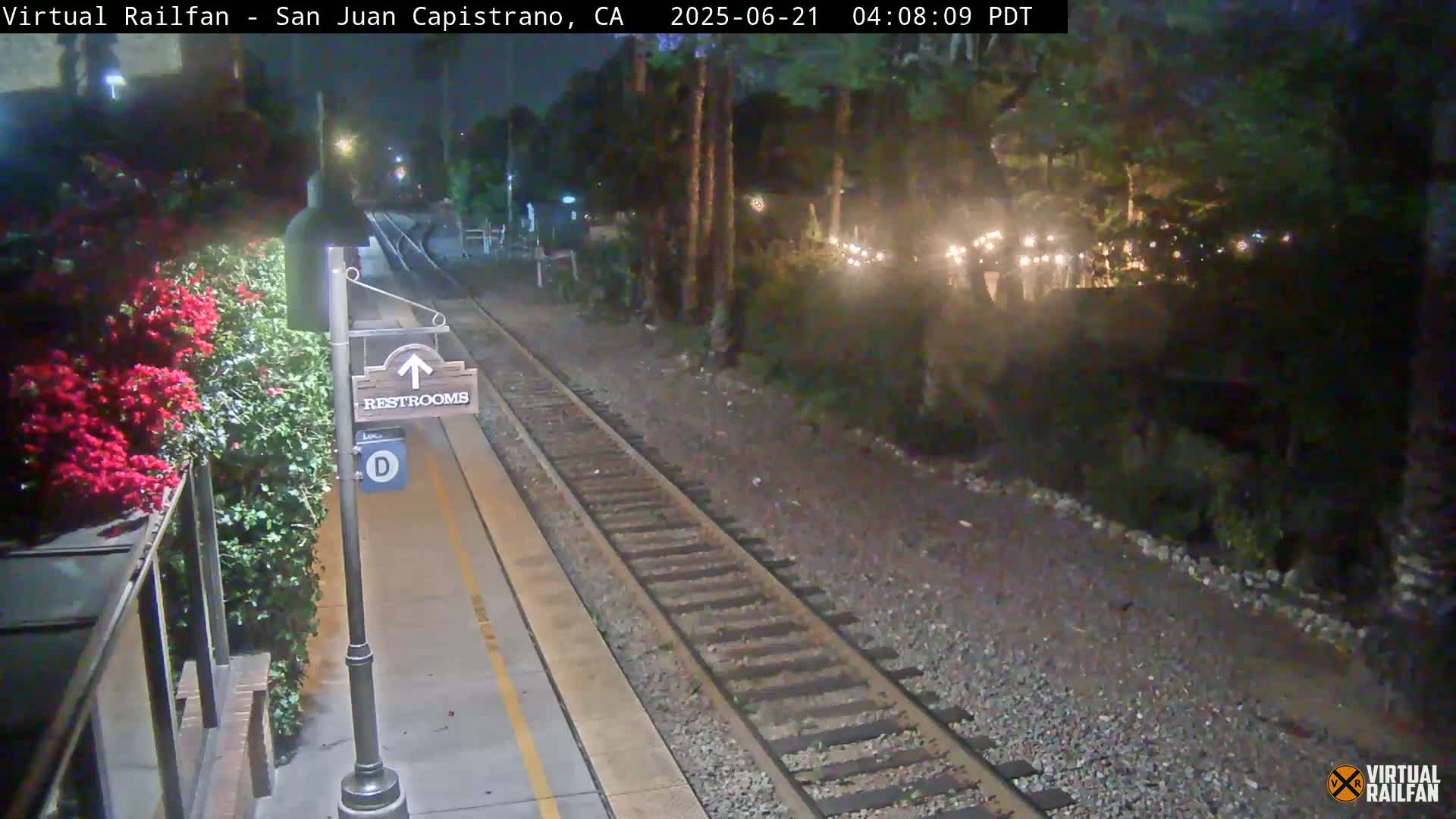 A nighttime view of a train platform with a restroom sign and vibrant red flowers beside a railway track.
