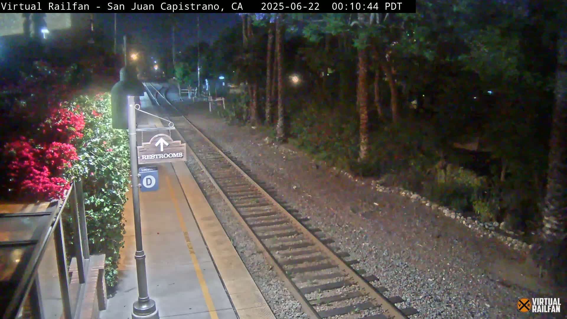 A nighttime view of a train platform with a restroom sign, lush greenery, and train tracks.
