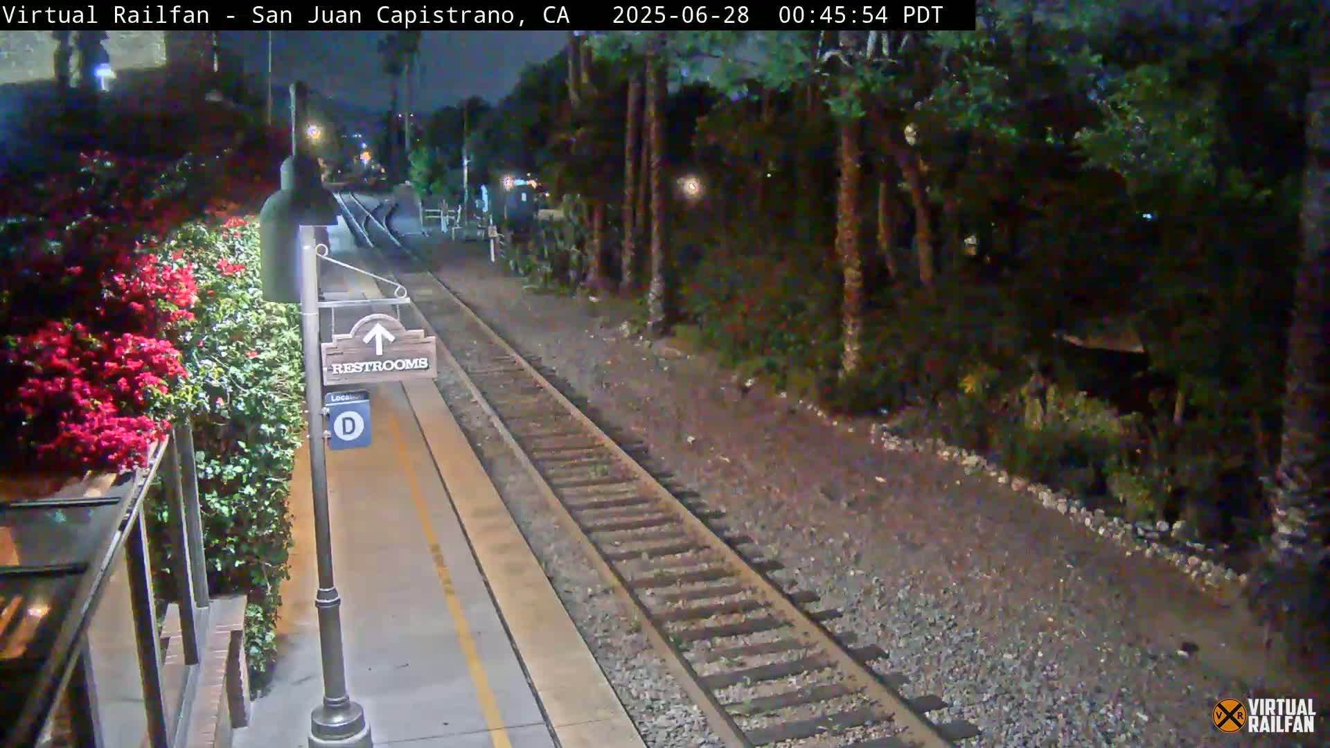 A nighttime view of a train platform with a restroom sign, alongside a railway track bordered by lush vegetation.
