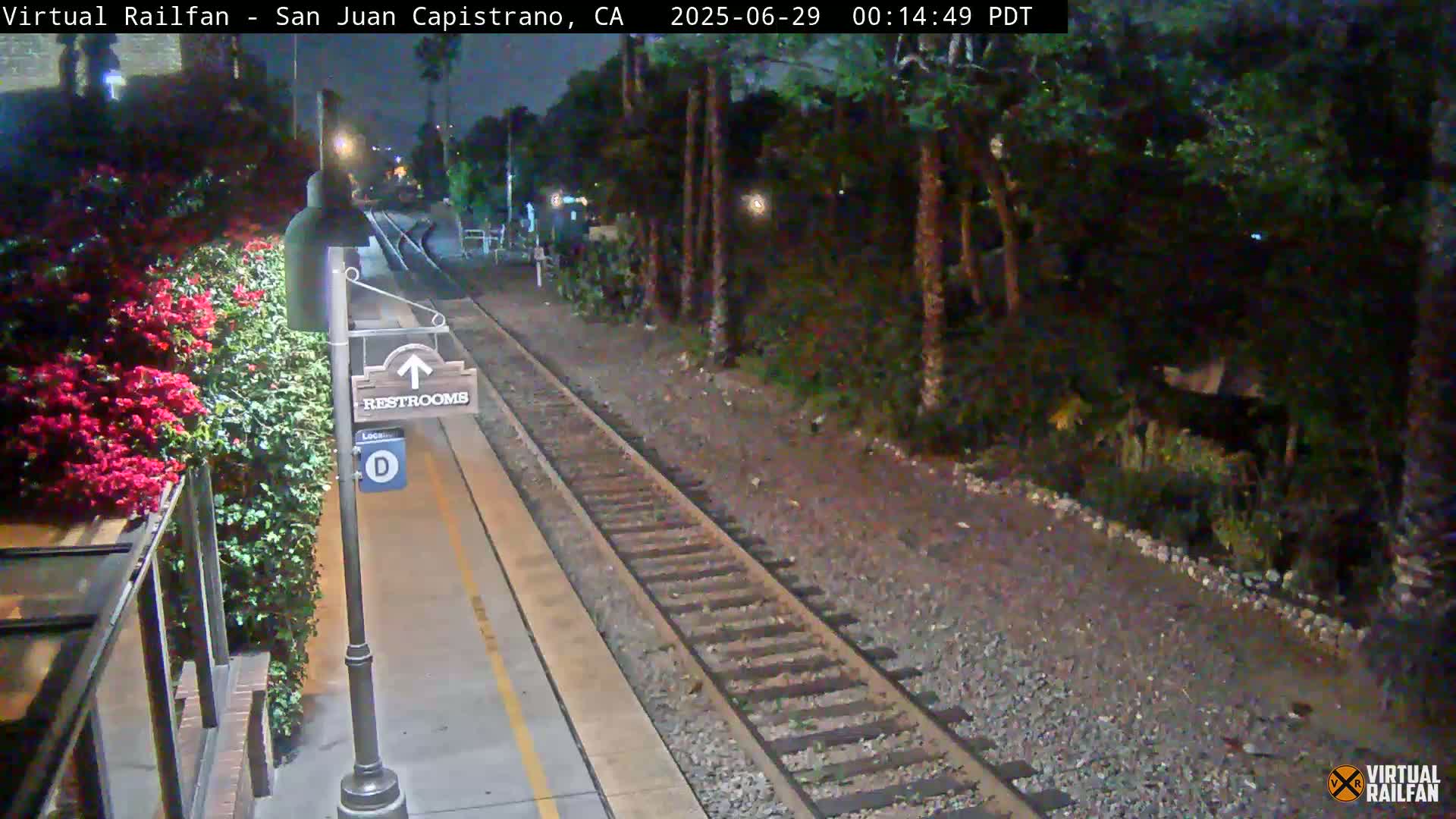 A nighttime view of a train platform with a restroom sign, railroad tracks, and lush greenery bordering the platform.