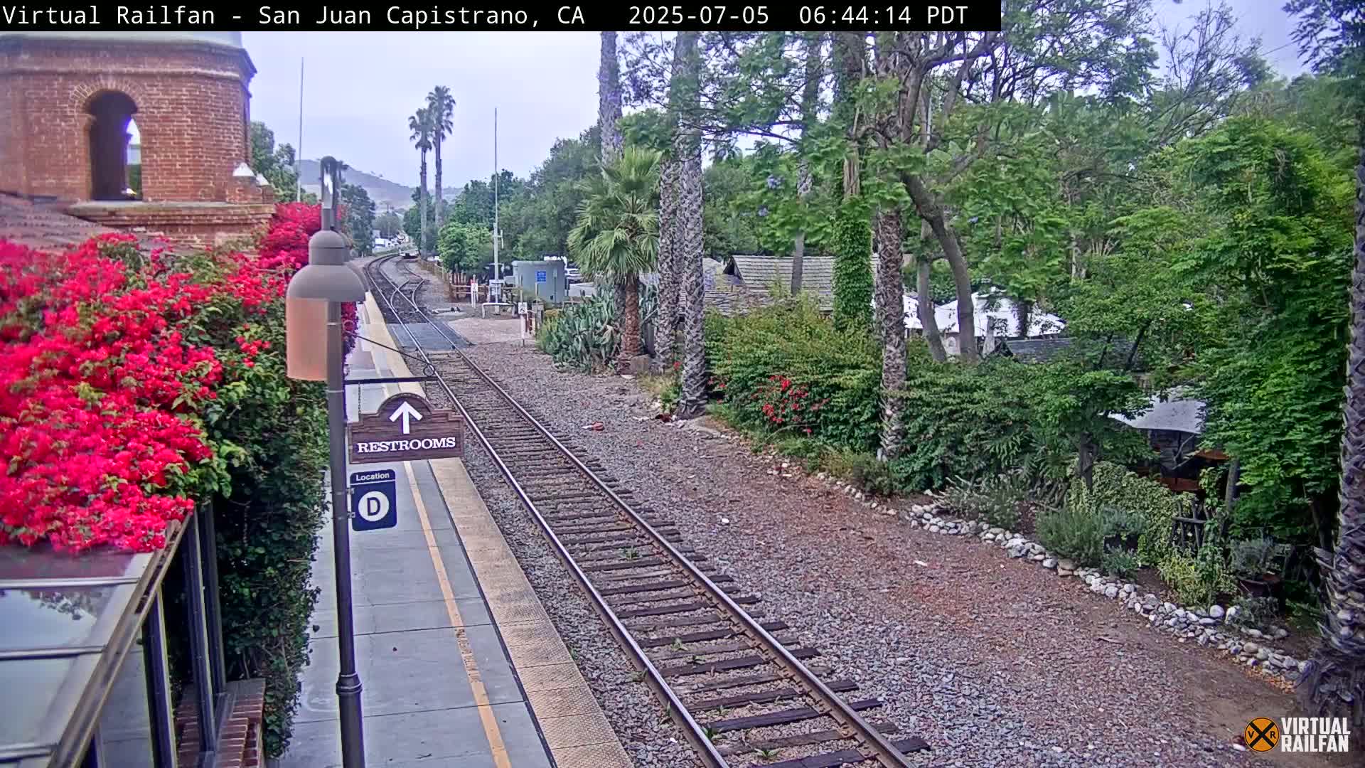 A train platform with a restroom sign, vibrant bougainvillea, and train tracks extends into a lush, tree-lined area under an overcast sky.