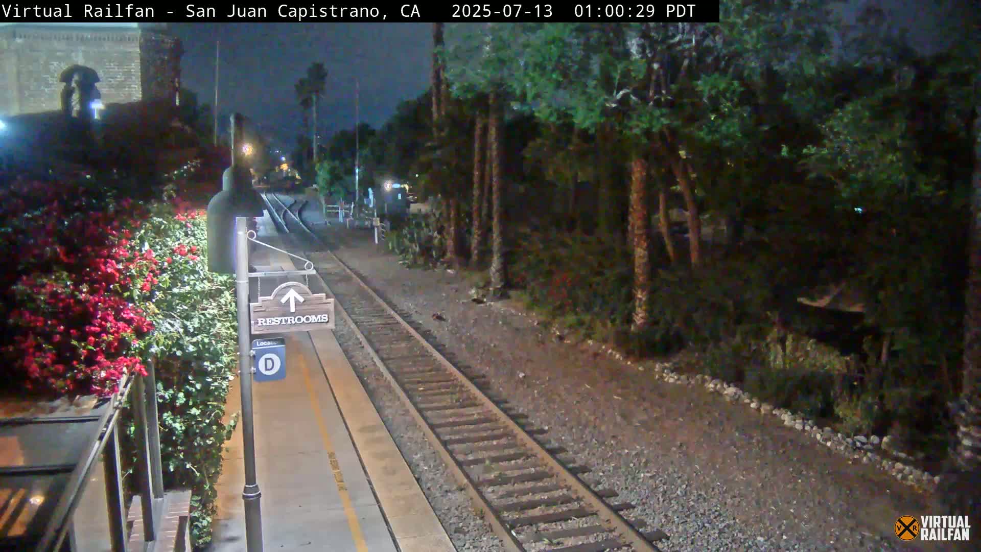 A nighttime view of a train station platform with a restroom sign, lush greenery, and train tracks extending into the darkness.