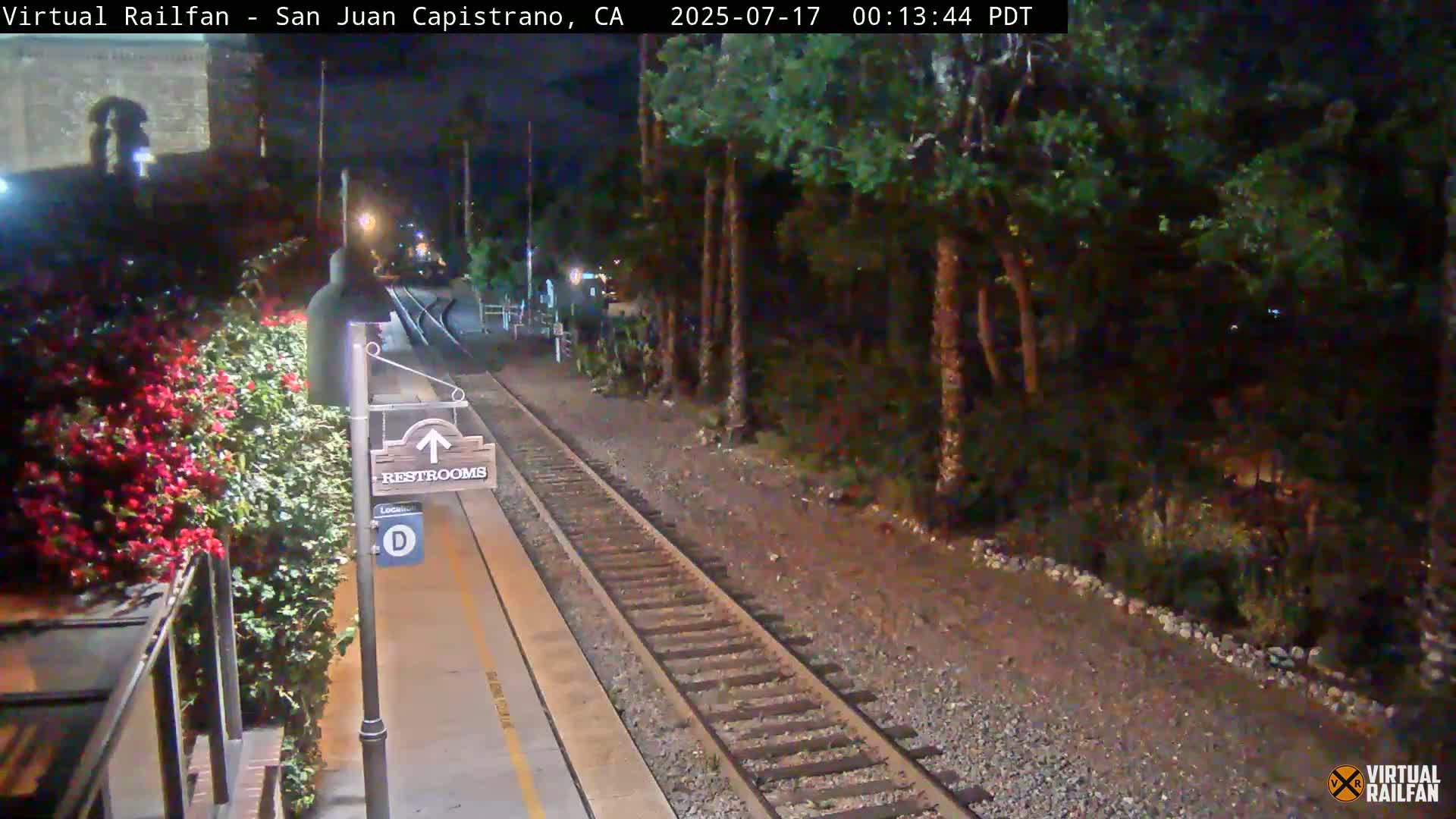 A nighttime view of a train platform with a restroom sign, blooming flowers, and train tracks extending into a dark, wooded area.