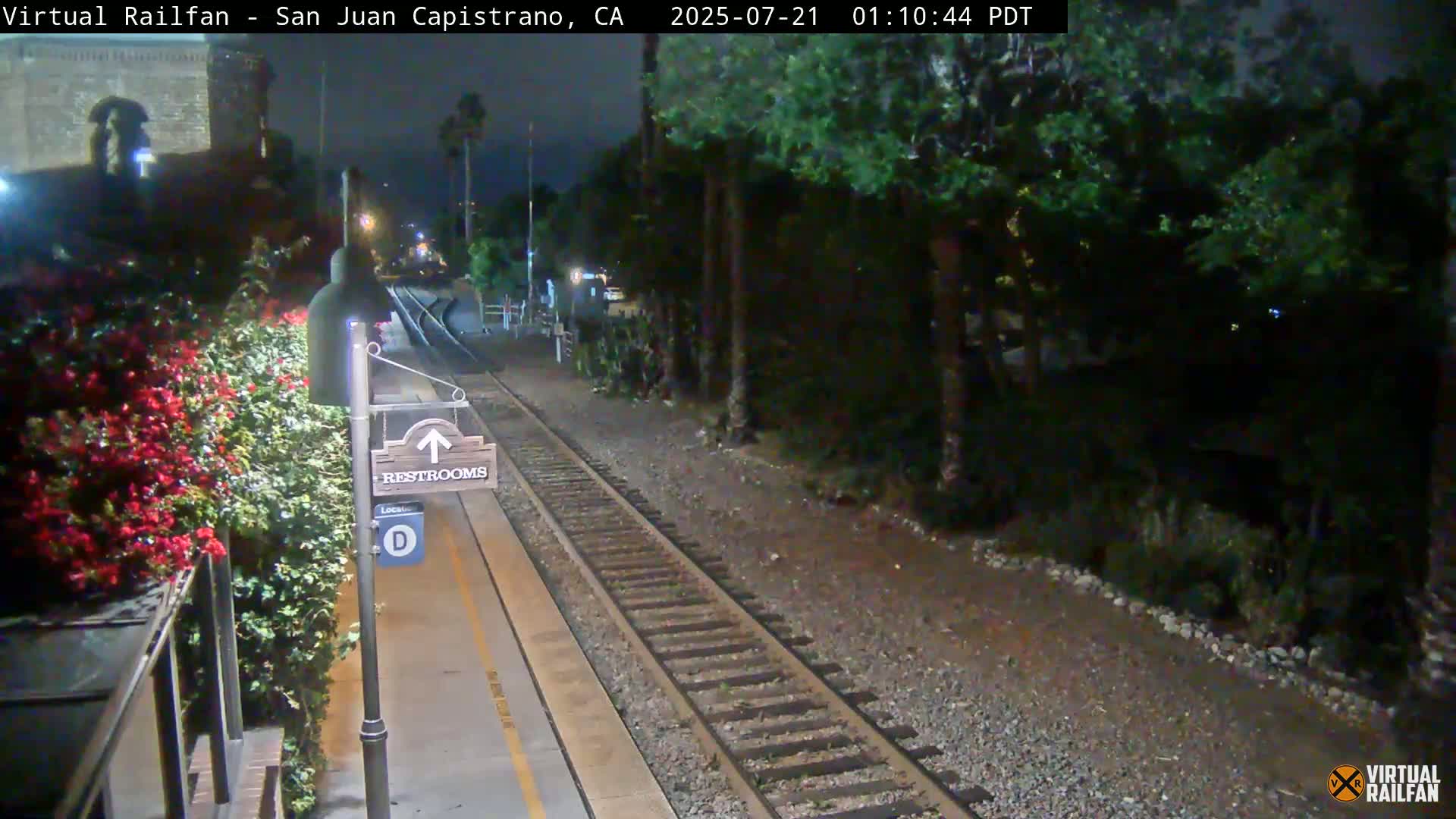 A nighttime view of a train platform with a restroom sign, railroad tracks, and lush vegetation alongside.